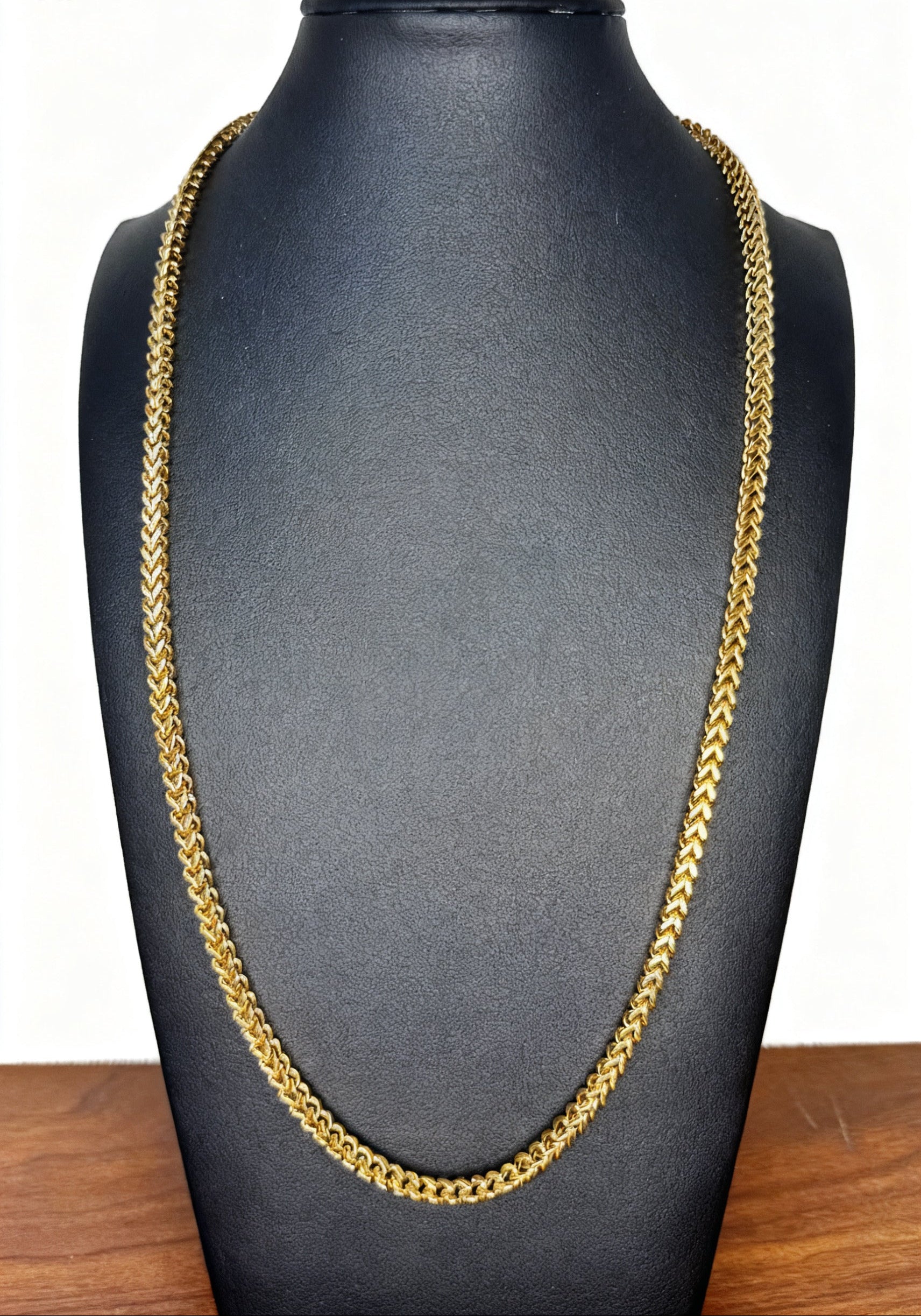 A gold-toned Franco chain necklace displayed on a black jewelry bust against a white background with a wooden surface visible at the bottom.