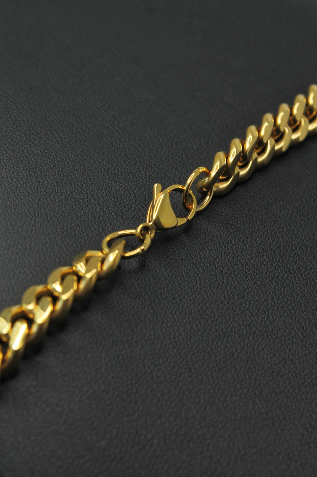 A close-up of a gold Curb link chain necklace with a lobster clasp, resting on a textured black surface.