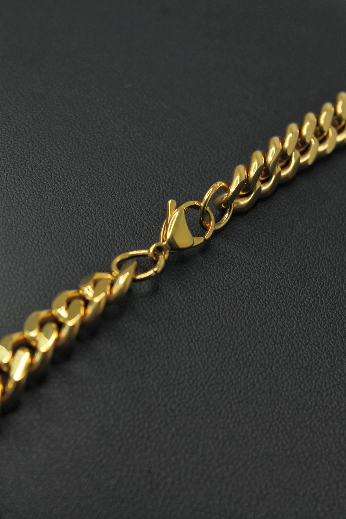 A close-up of a gold Curb link chain necklace with a lobster clasp, resting on a textured black surface.