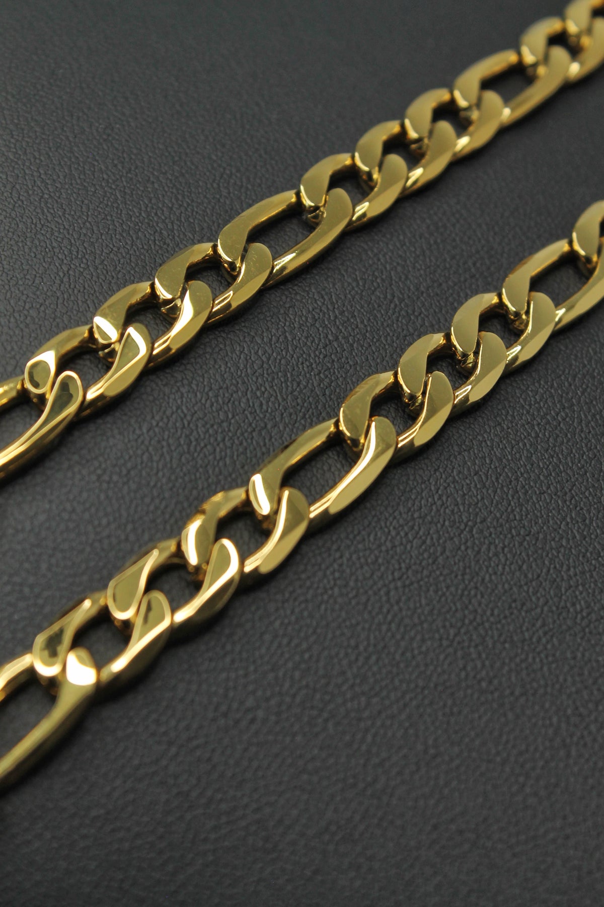 A close-up of a shiny gold-tone Figaro link chain draped diagonally across a dark textured surface.