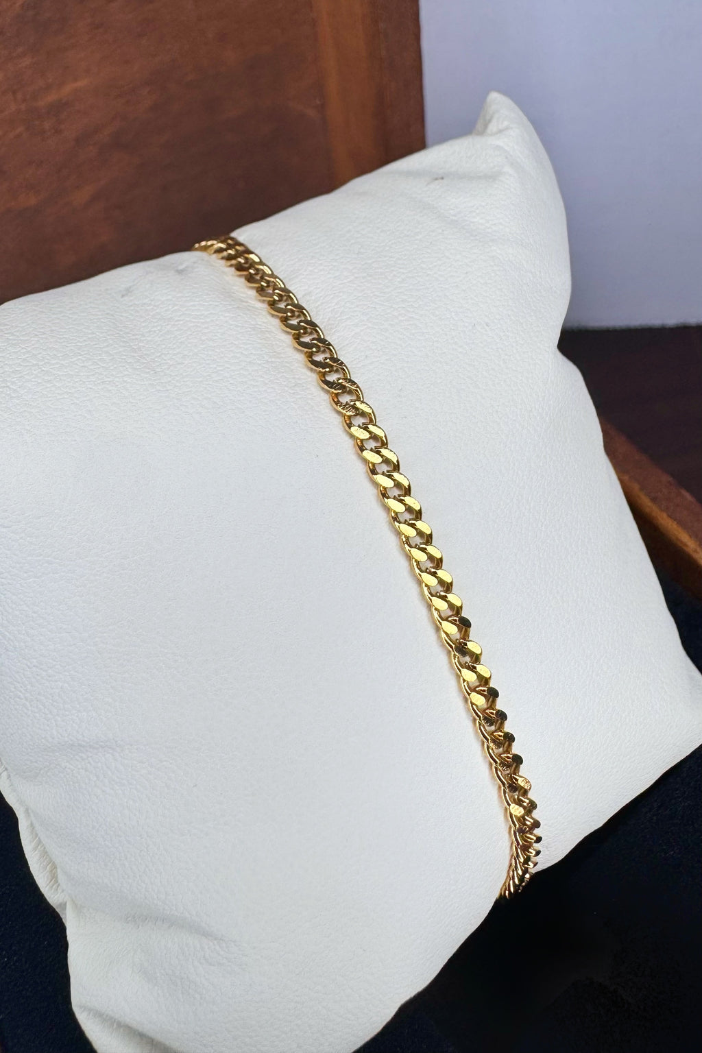 A gold-toned Curb link chain bracelet draped diagonally across a white textured pillow, with a dark wooden surface in the background.
