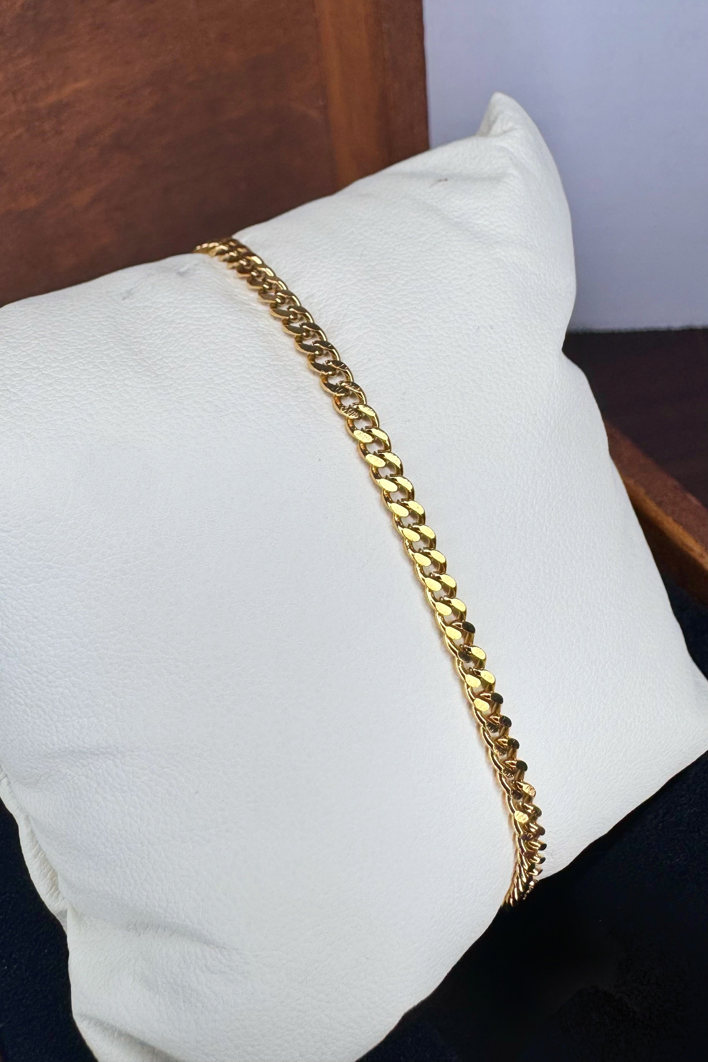 A gold-toned Curb link chain bracelet draped diagonally across a white textured pillow, with a dark wooden surface in the background.