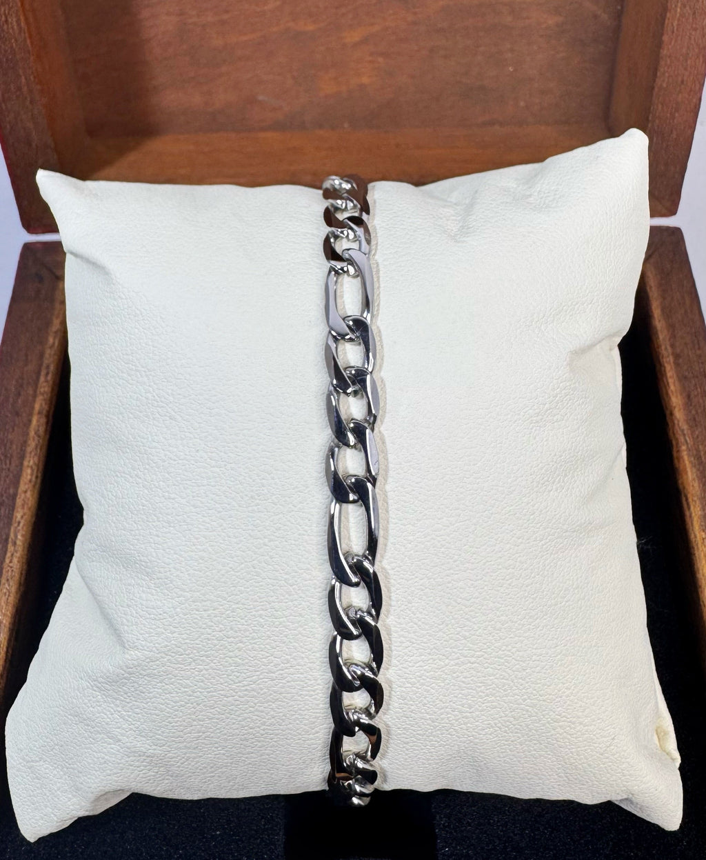 A silver-toned chunky figaro chain link bracelet draped vertically down the center of a white textured pillow, set inside a dark brown wooden box.
