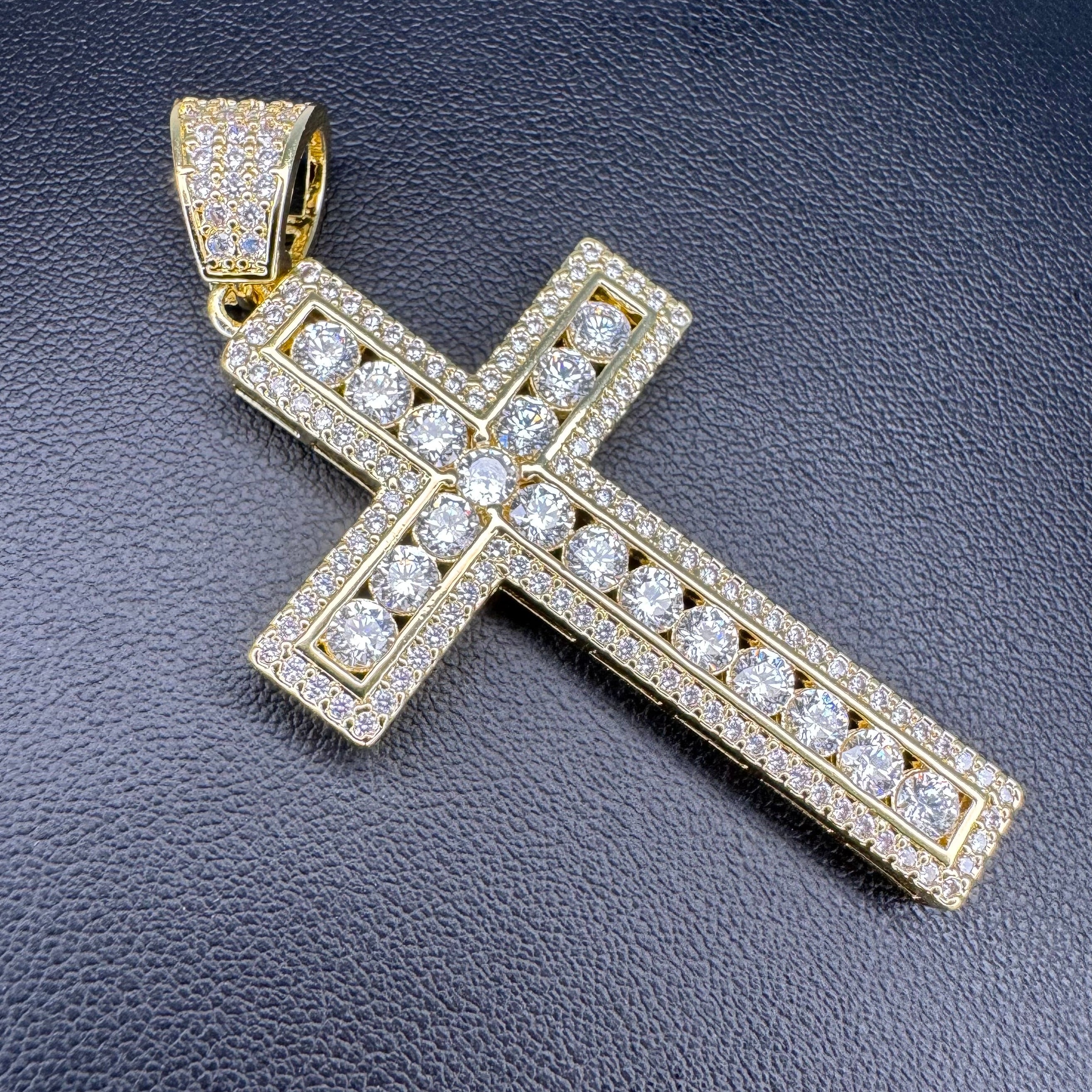 A gold cross pendant encrusted with clear cubic zirconia stones, displayed on a textured black background.
