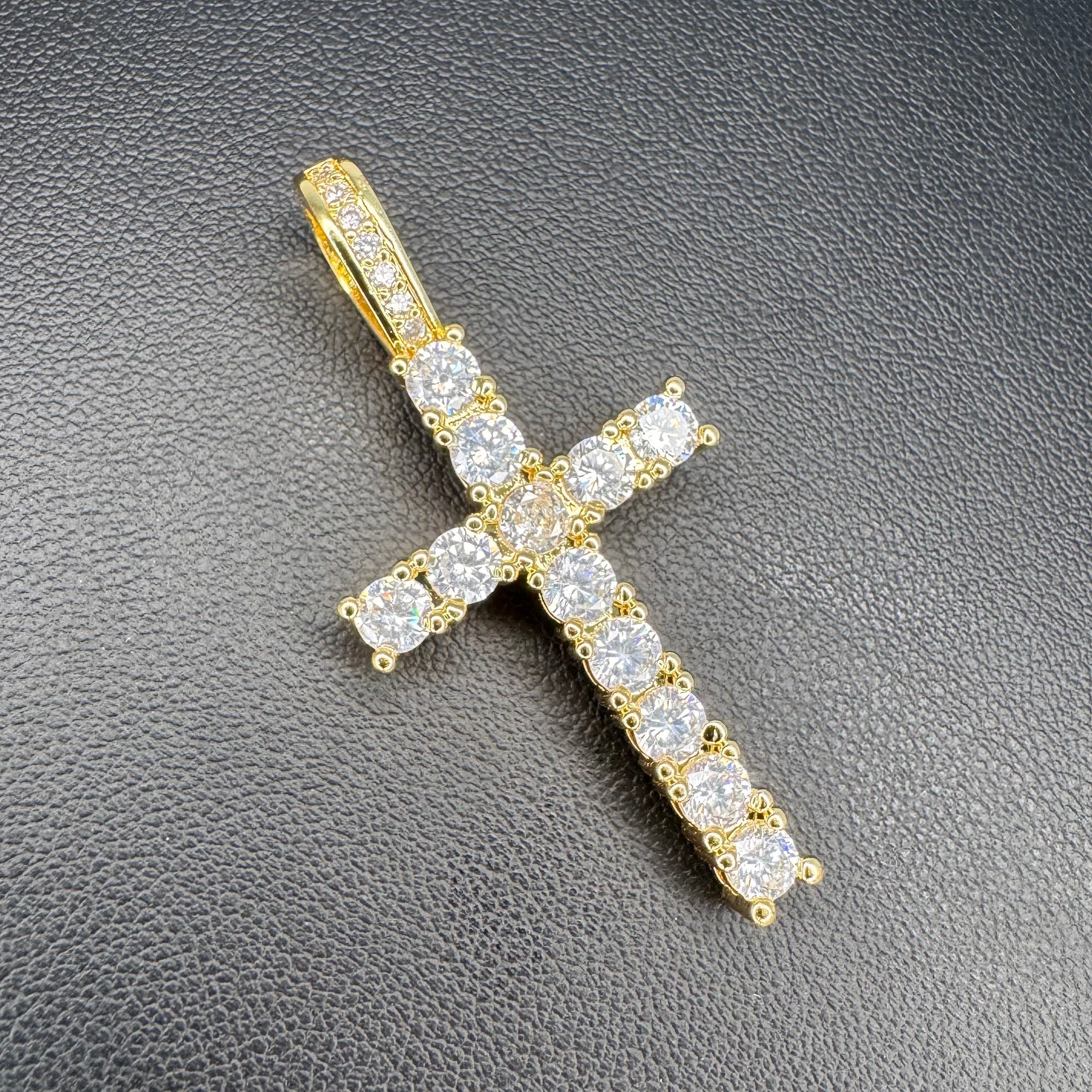 A yellow gold cross pendant encrusted with clear round-cut gemstones, displayed on a textured black surface.