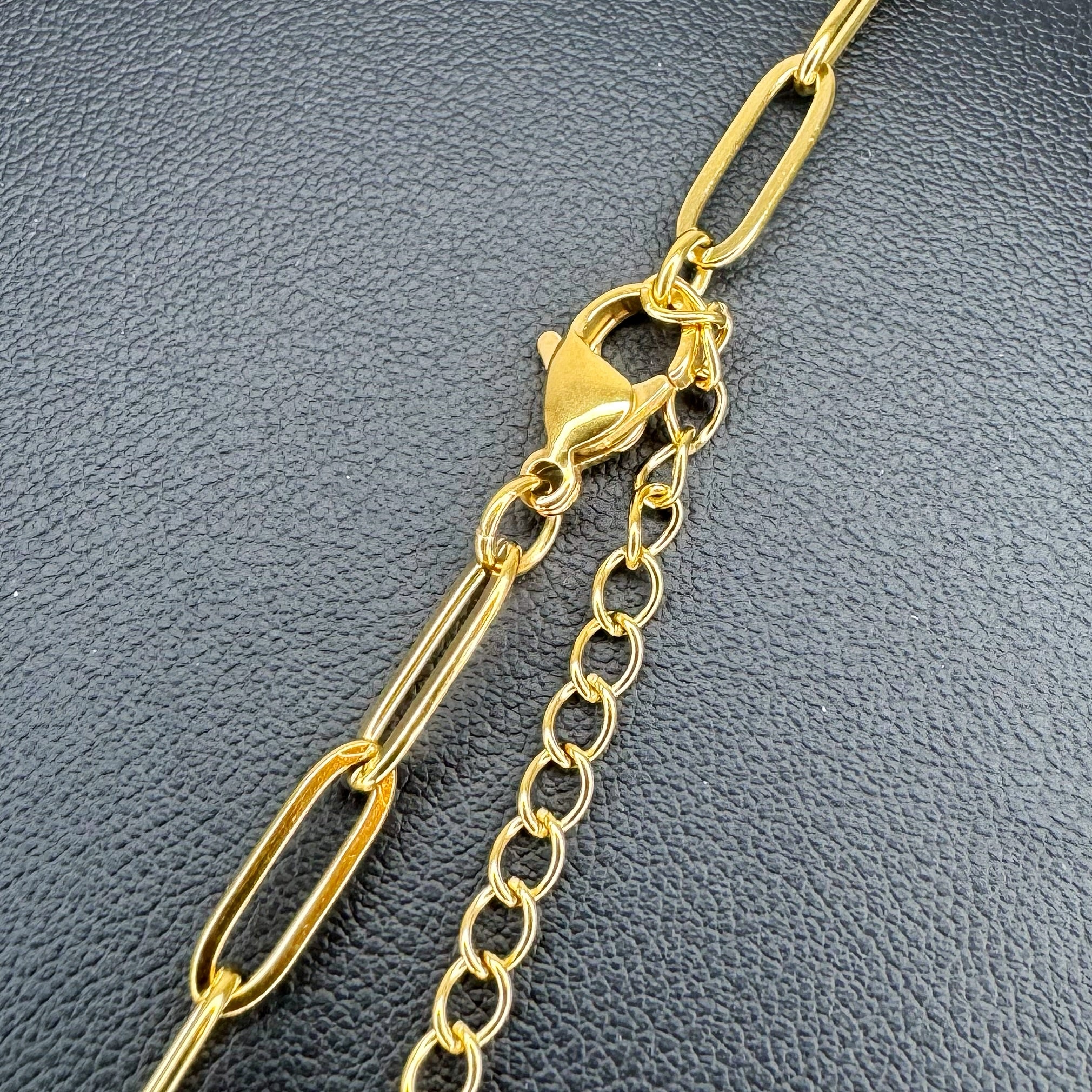 A close-up of a gold-tone chain accessory featuring a paperclip link design, a lobster clasp, and an extension chain, displayed on a textured dark gray surface.