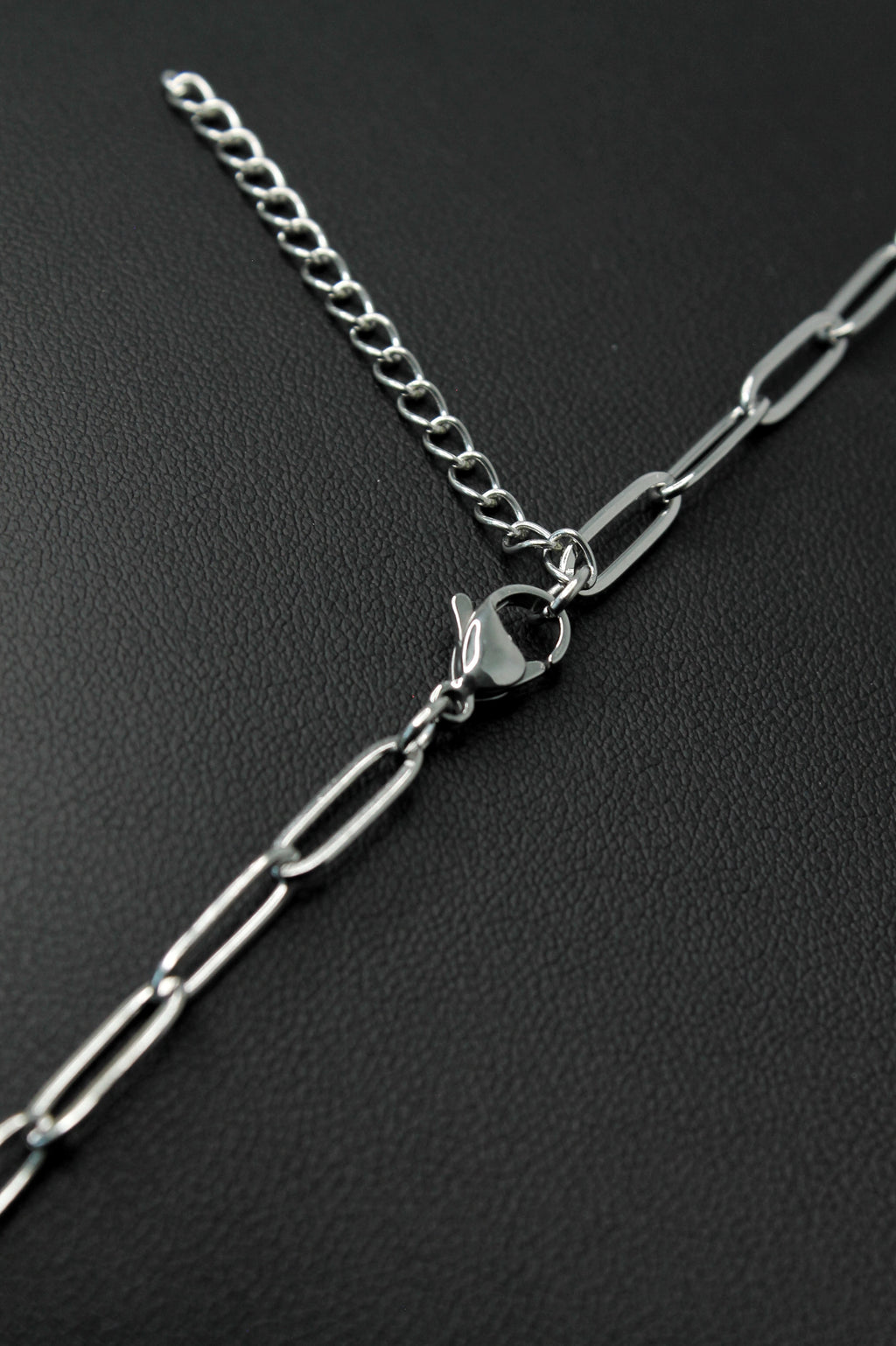 A close-up of a silver chain extender with a lobster clasp, featuring a fine spring ring chain and a thicker paperclip-style chain, displayed on a textured black background.