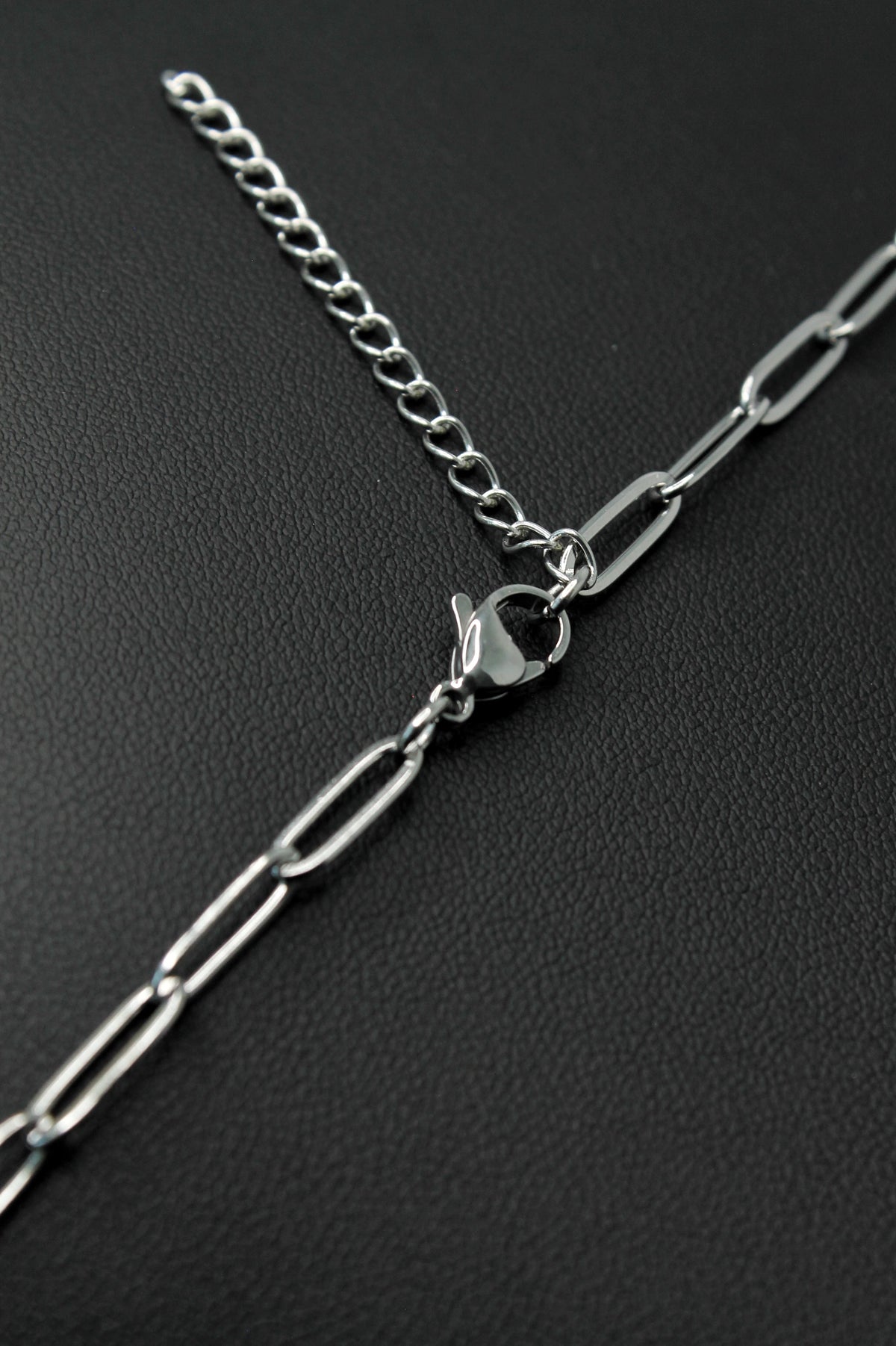 A close-up of a silver chain extender with a lobster clasp, featuring a fine spring ring chain and a thicker paperclip-style chain, displayed on a textured black background.