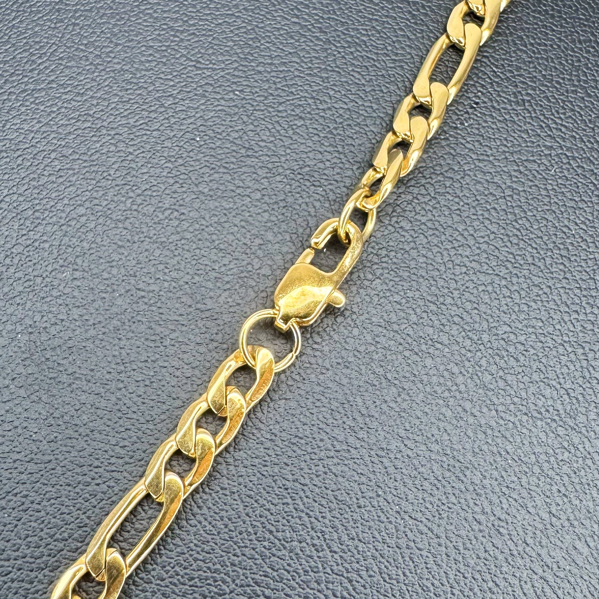 A close-up of a gold-toned metal chain Figaro link necklace with a lobster clasp, laid diagonally on a textured dark gray surface.