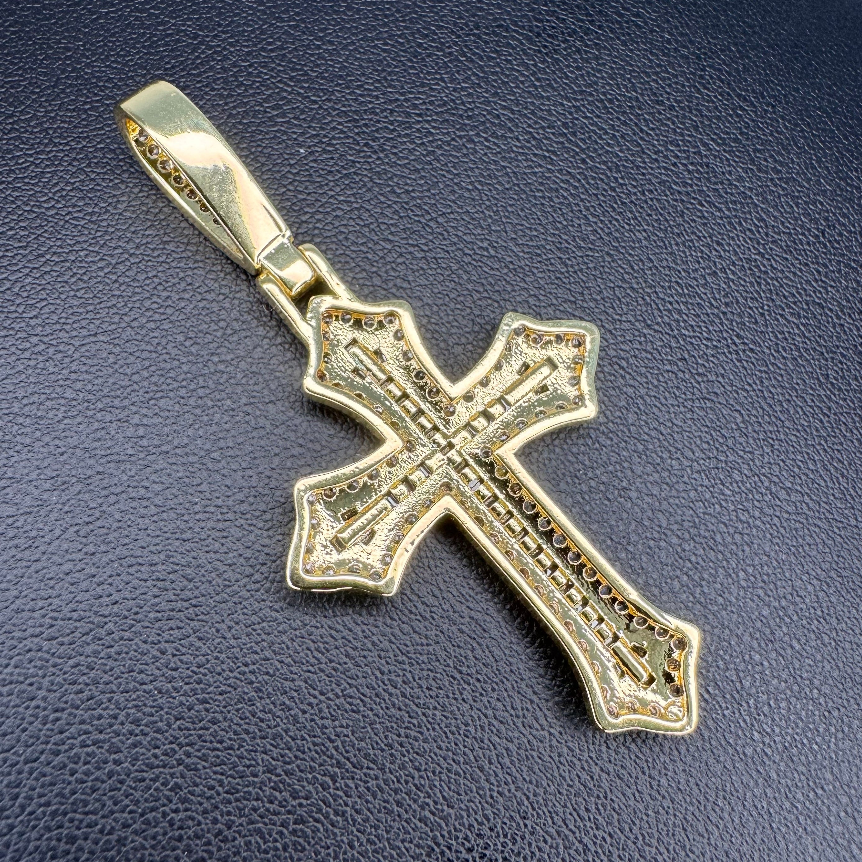 A gold cross pendant with intricate filigree detailing, displayed on a textured dark blue surface.