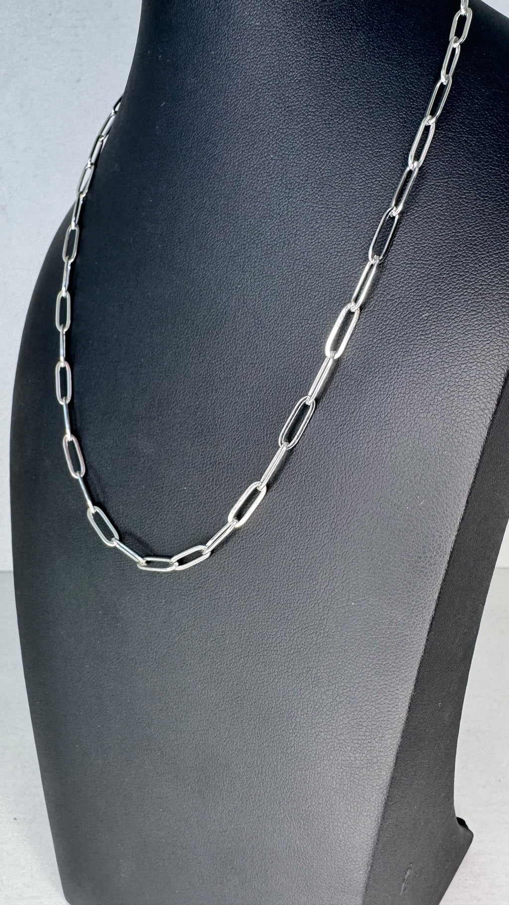 A silver paperclip chain necklace displayed on a black jewelry bust against a light gray background.