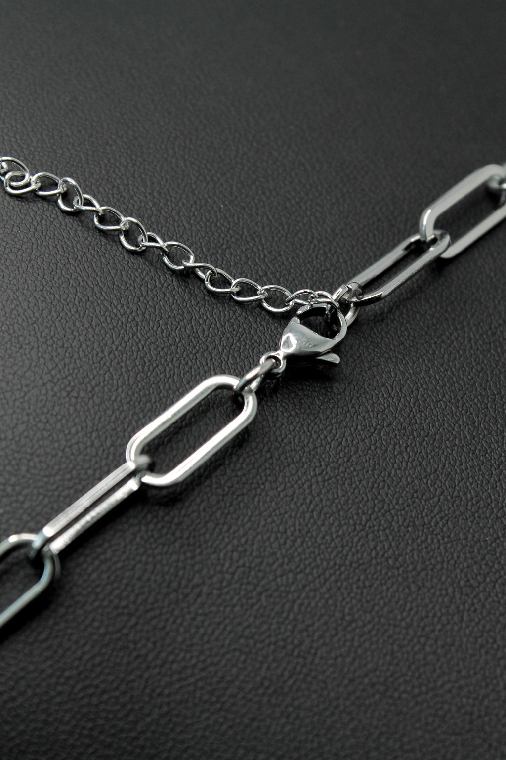 A close-up of a silver paperclip chain necklace with a lobster clasp, resting on a textured black surface.