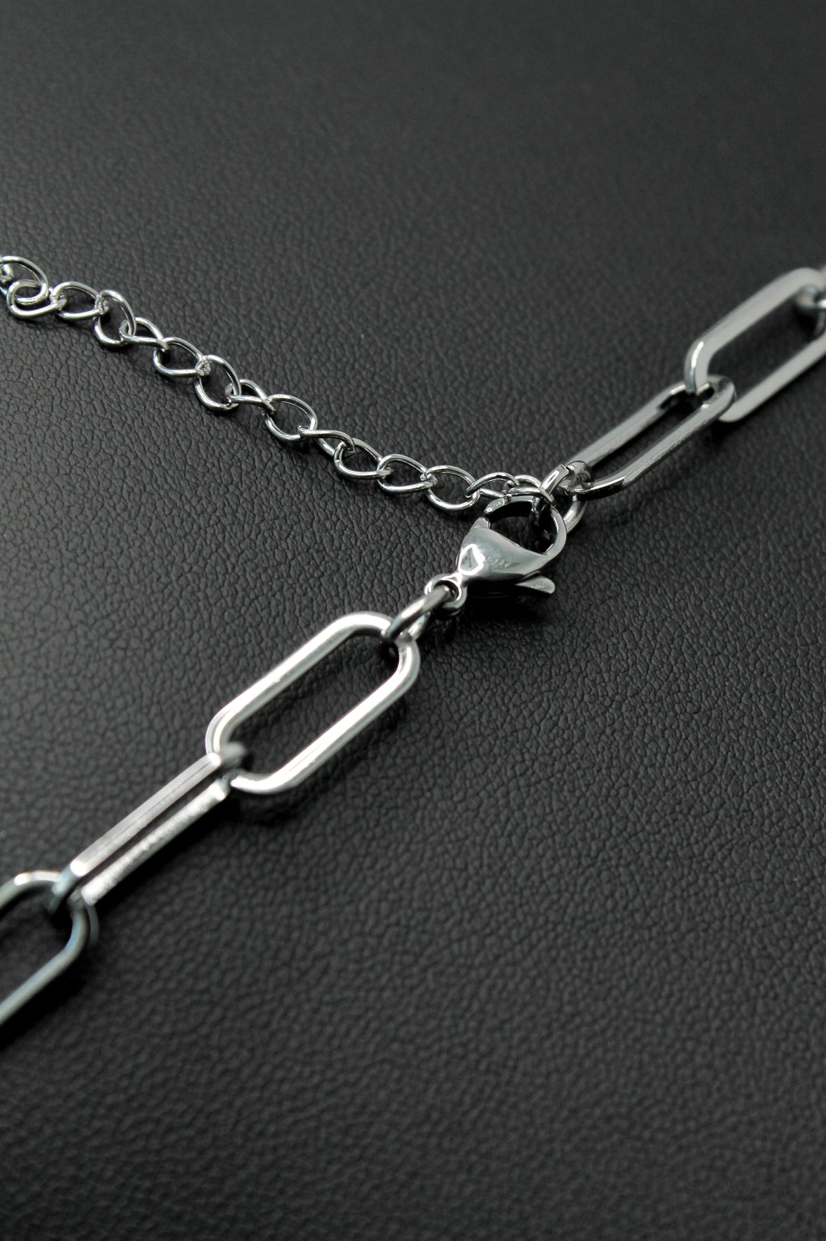 A close-up of a silver paperclip chain necklace with a lobster clasp, resting on a textured black surface.