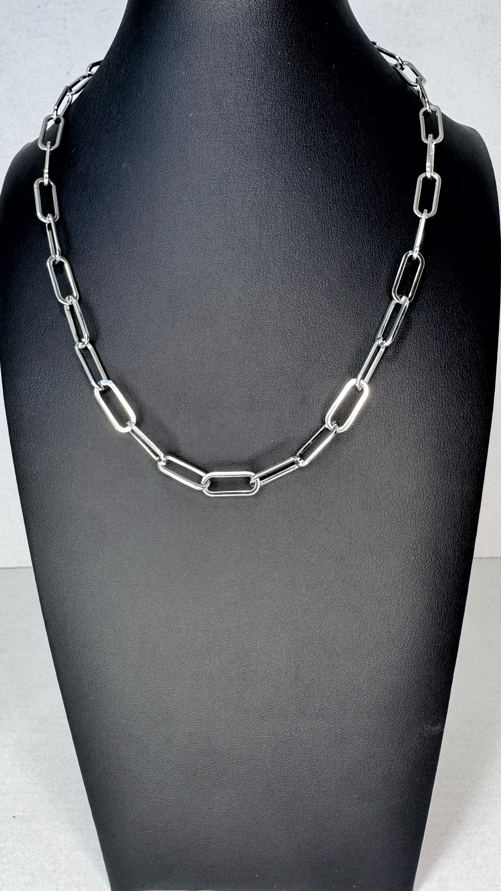 A silver paperclip chain necklace displayed on a black jewelry bust against a light gray background.