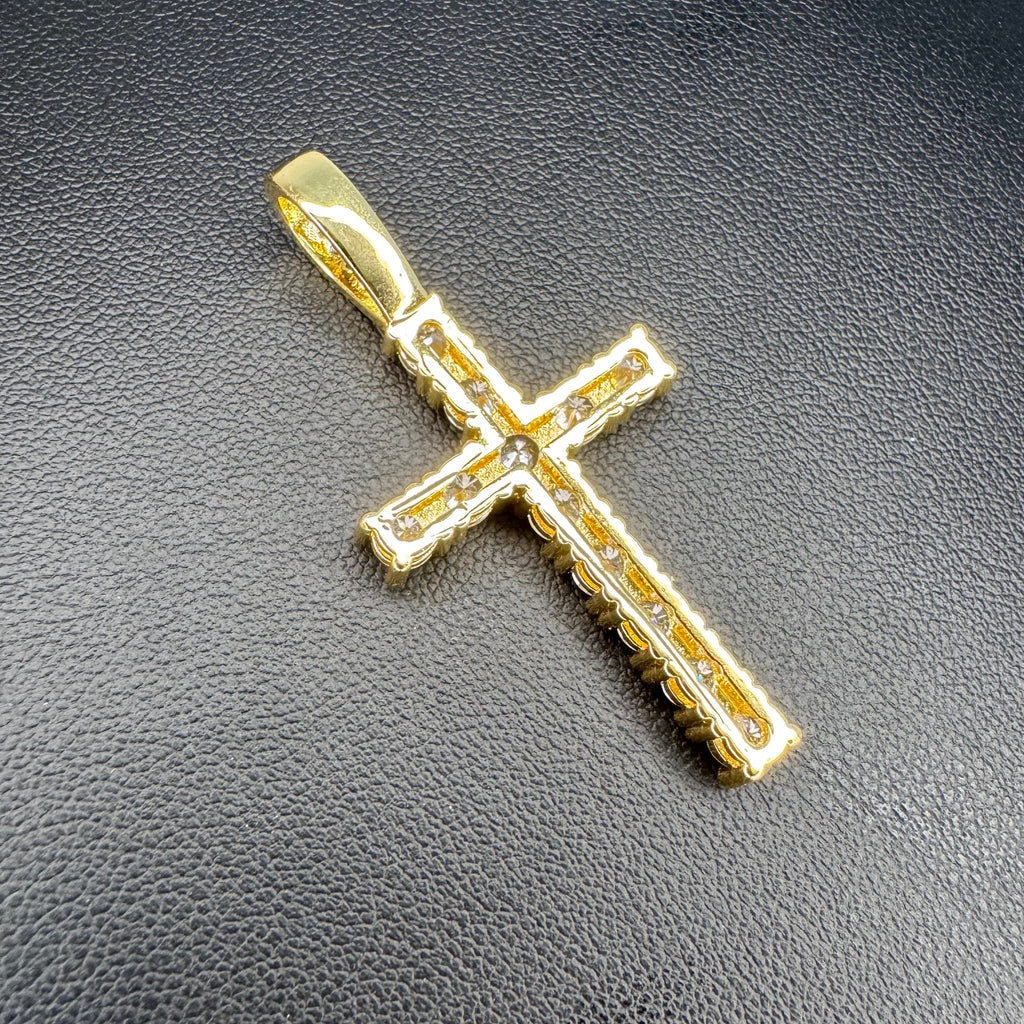 A gold cross pendant with clear gemstones set along the edges, displayed on a textured black surface.