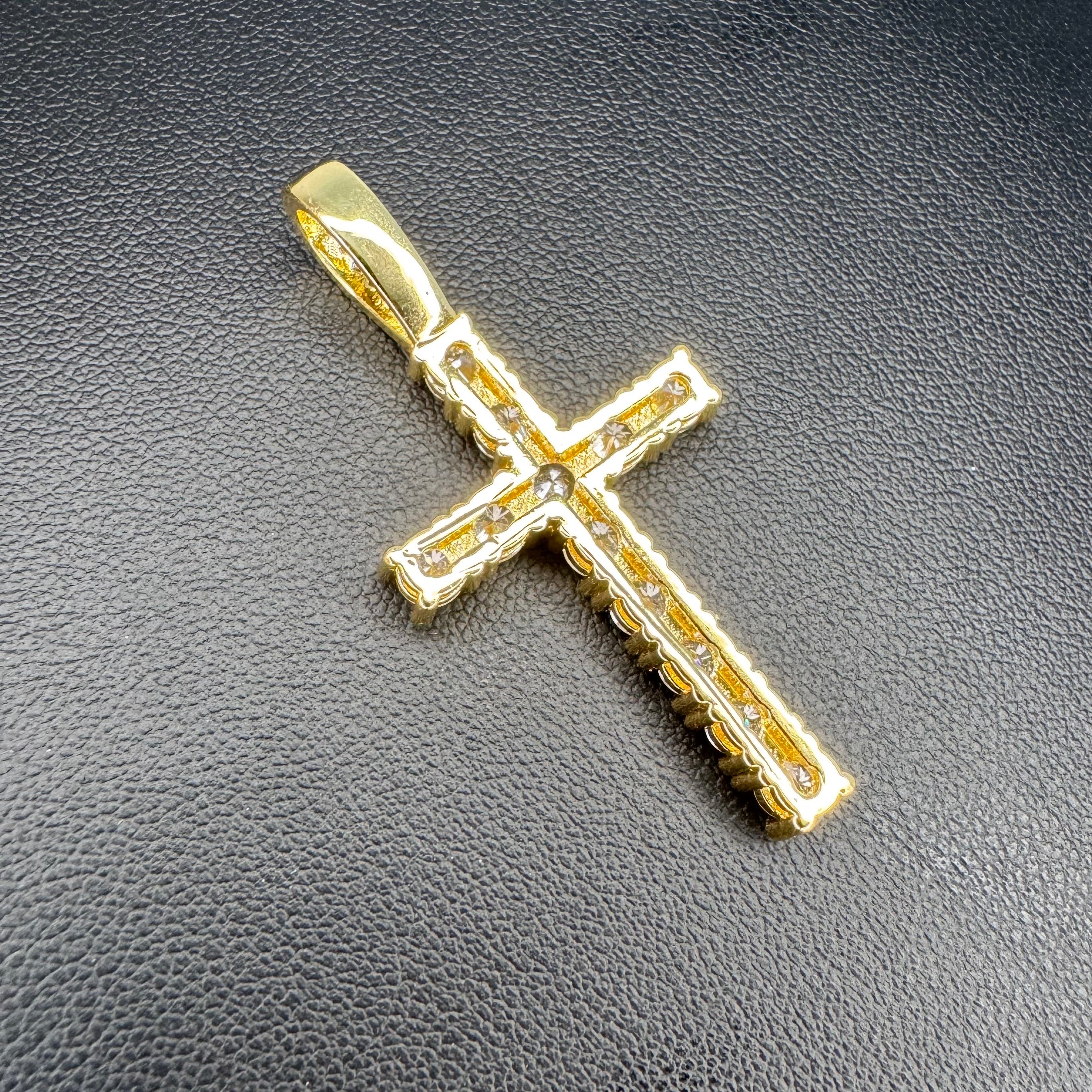 A gold cross pendant with clear gemstones set along the edges, displayed on a textured black surface.