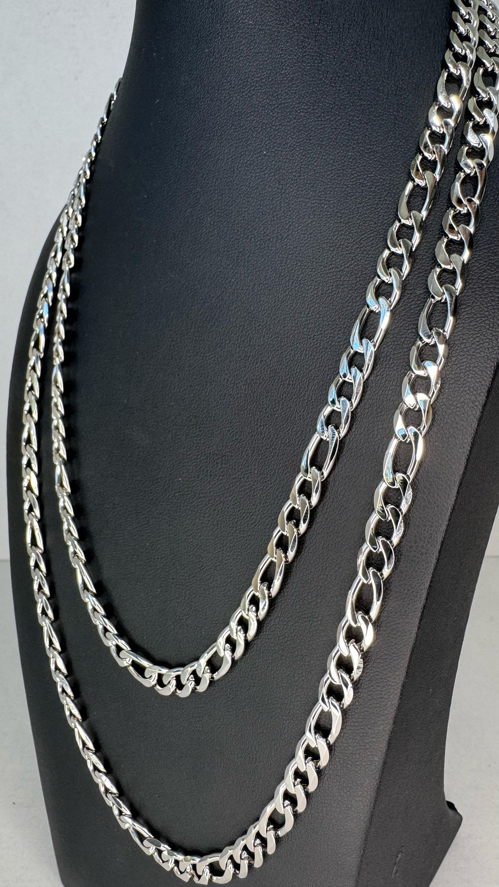 Two silver-toned Figaro chain necklaces displayed on a black mannequin bust against a light gray background.