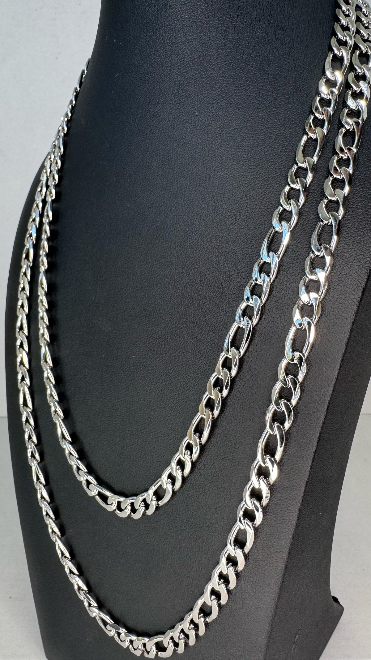 Two silver-toned Figaro chain necklaces displayed on a black mannequin bust against a light gray background.