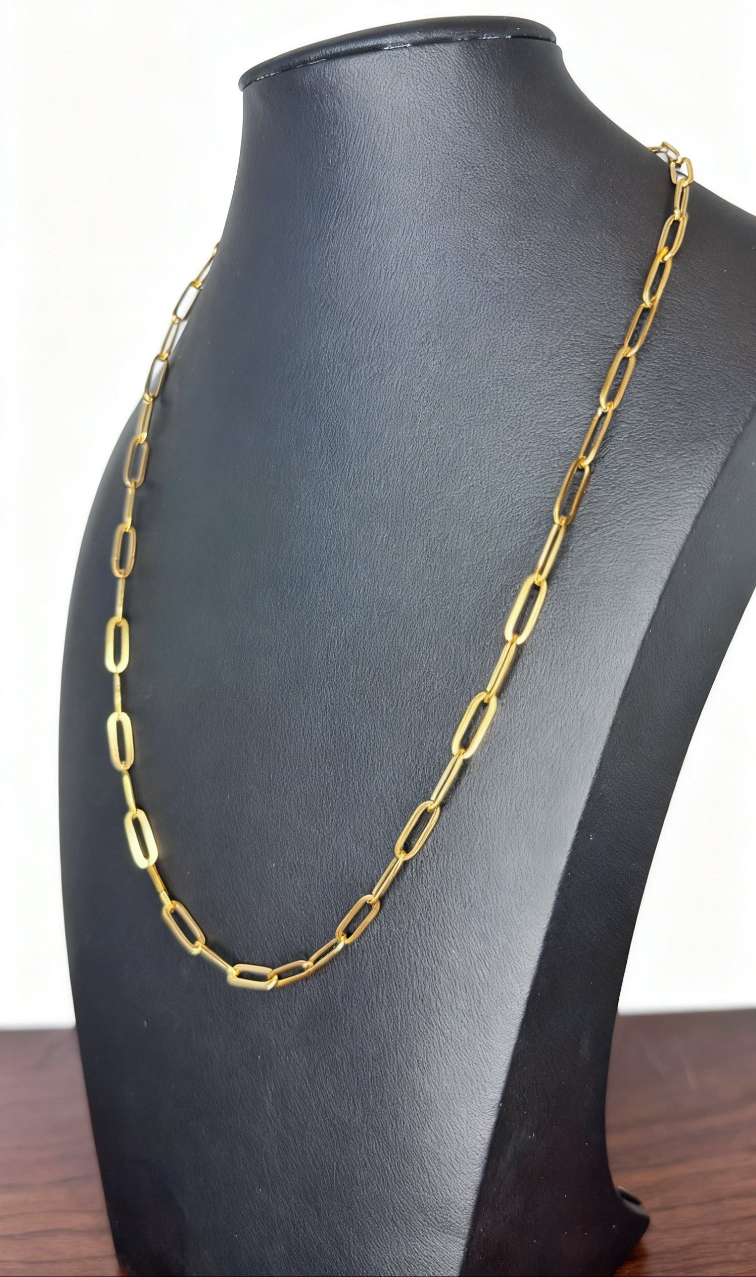A gold paperclip chain necklace displayed on a black jewelry bust against a white background.
