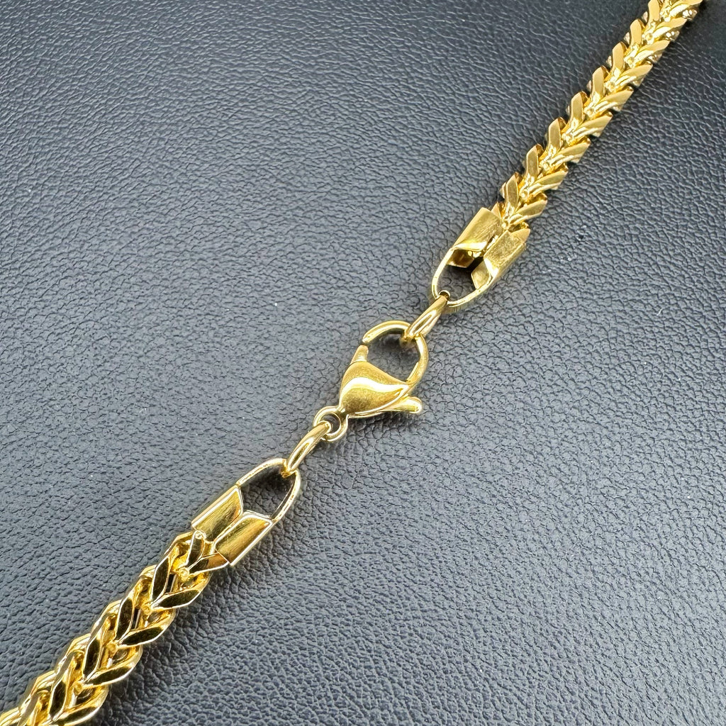 A close-up of a gold-toned Franco chain necklace with a textured wheat-like link pattern and a lobster clasp, laid diagonally on a dark gray textured surface.