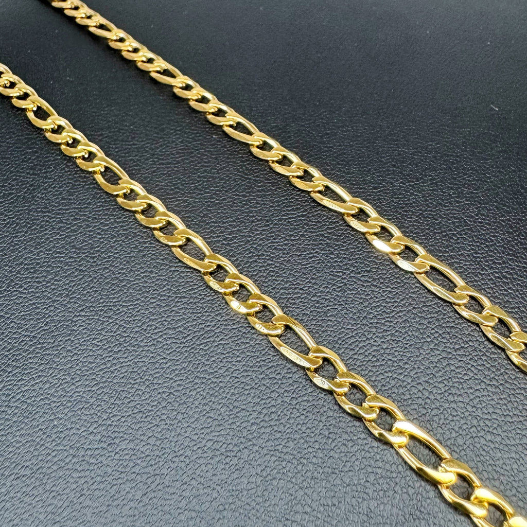A close-up of a polished yellow gold Figaro chain with elongated oval links, laid diagonally on a textured black surface.