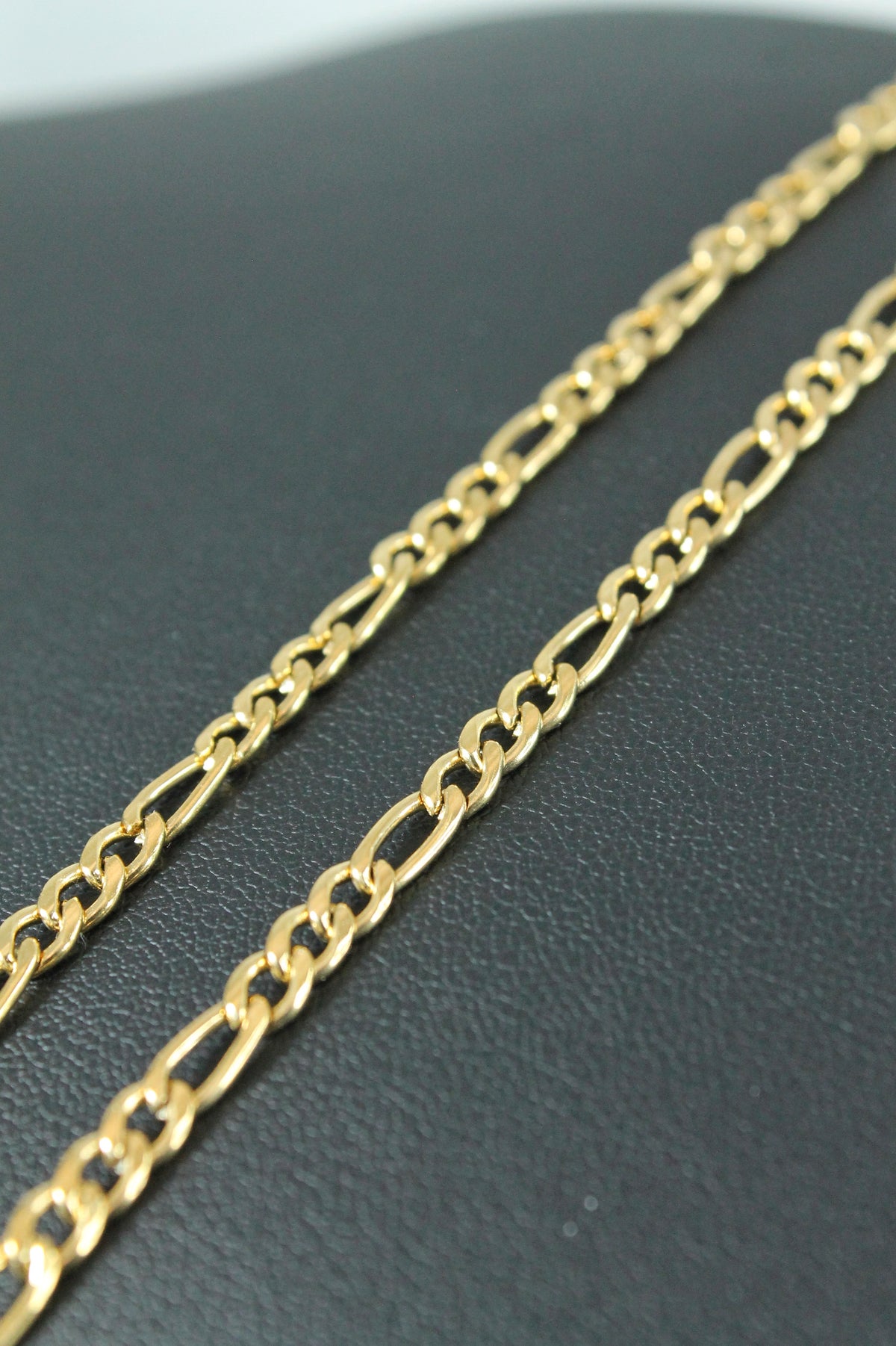 A close-up of a polished yellow gold Figaro chain necklace resting on a textured black surface.