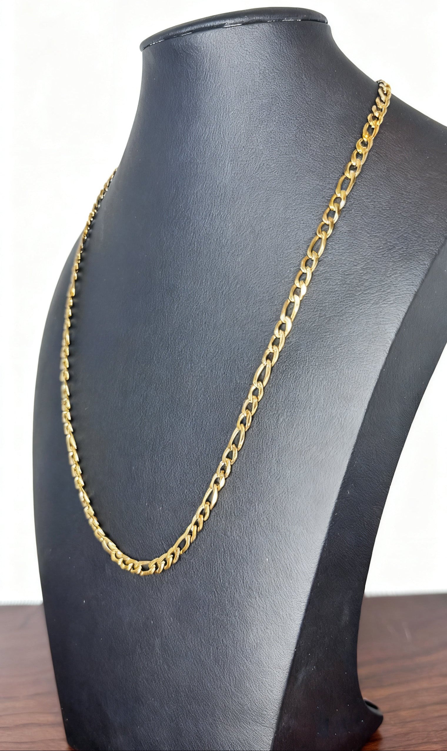 A gold-toned Figaro chain necklace displayed on a black jewelry bust against a white background.