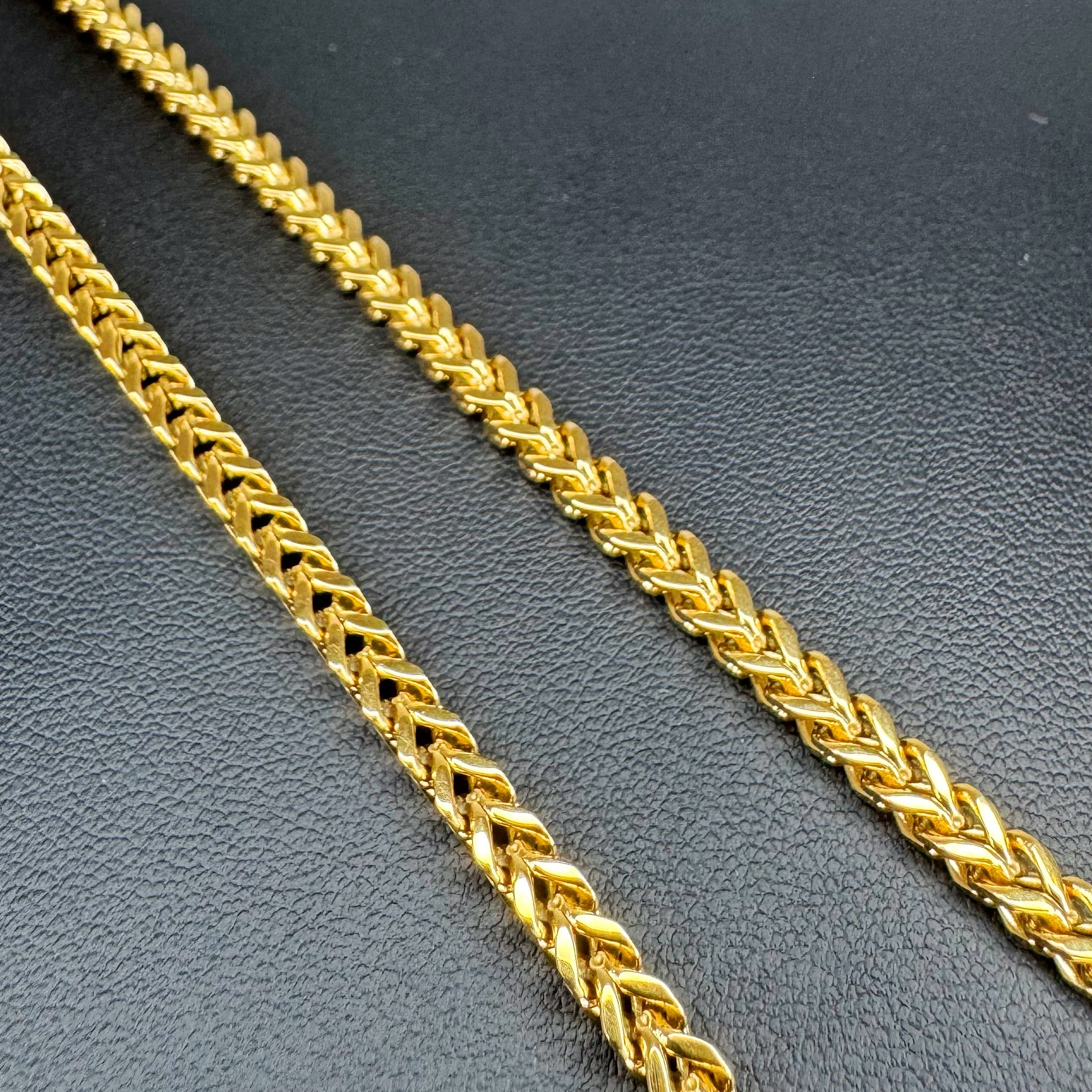 A close-up of a shiny gold Franco link chain laid diagonally on a dark textured surface.