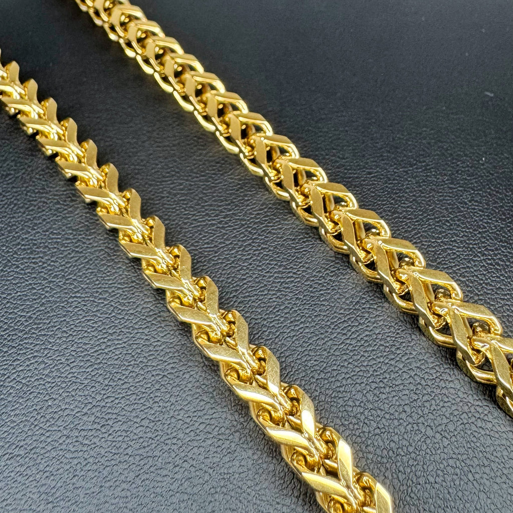 A close-up of a shiny gold chain necklace Franco link design, laid diagonally on a dark textured surface.