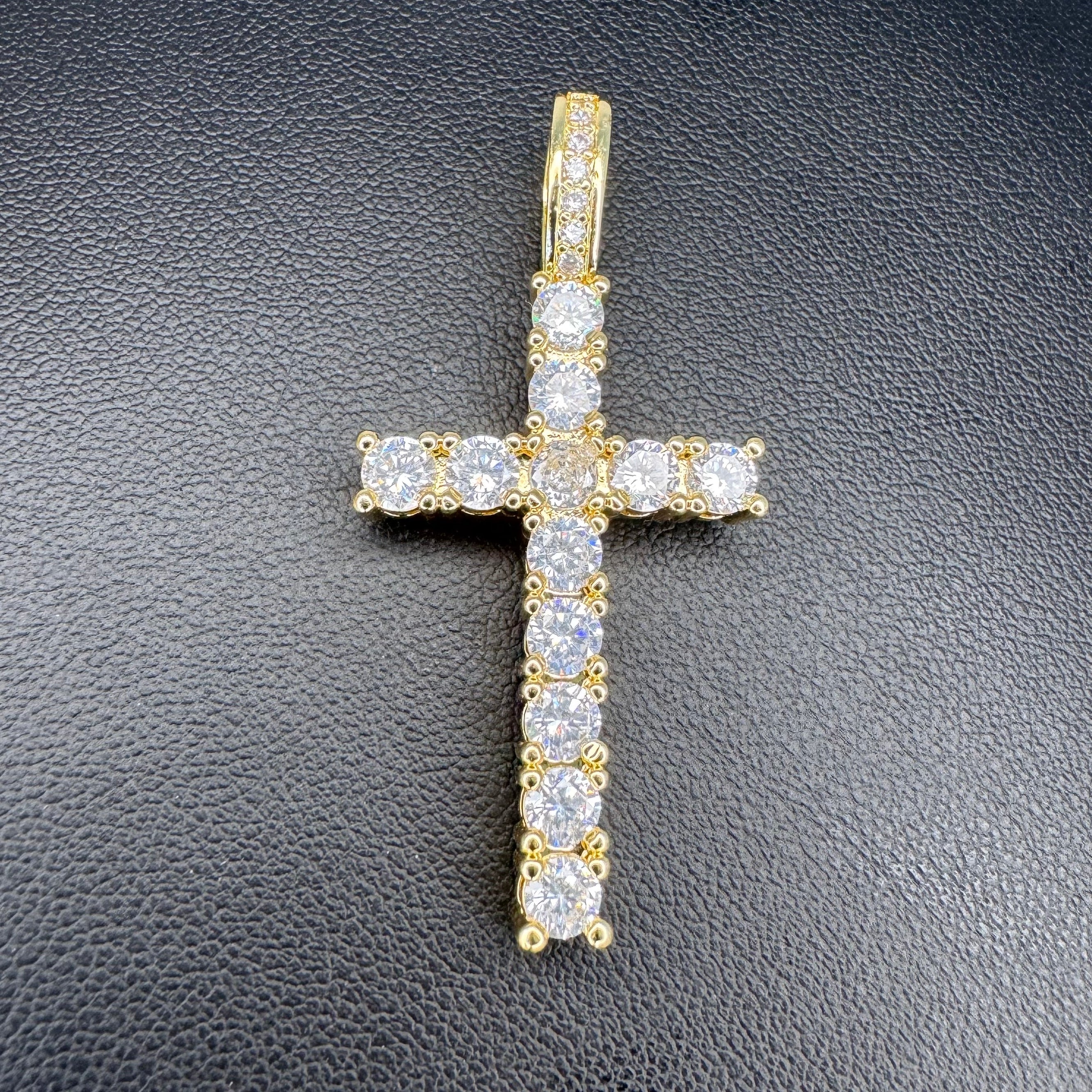 A yellow gold cross pendant adorned with clear cubic zirconia stones, displayed on a textured black surface.