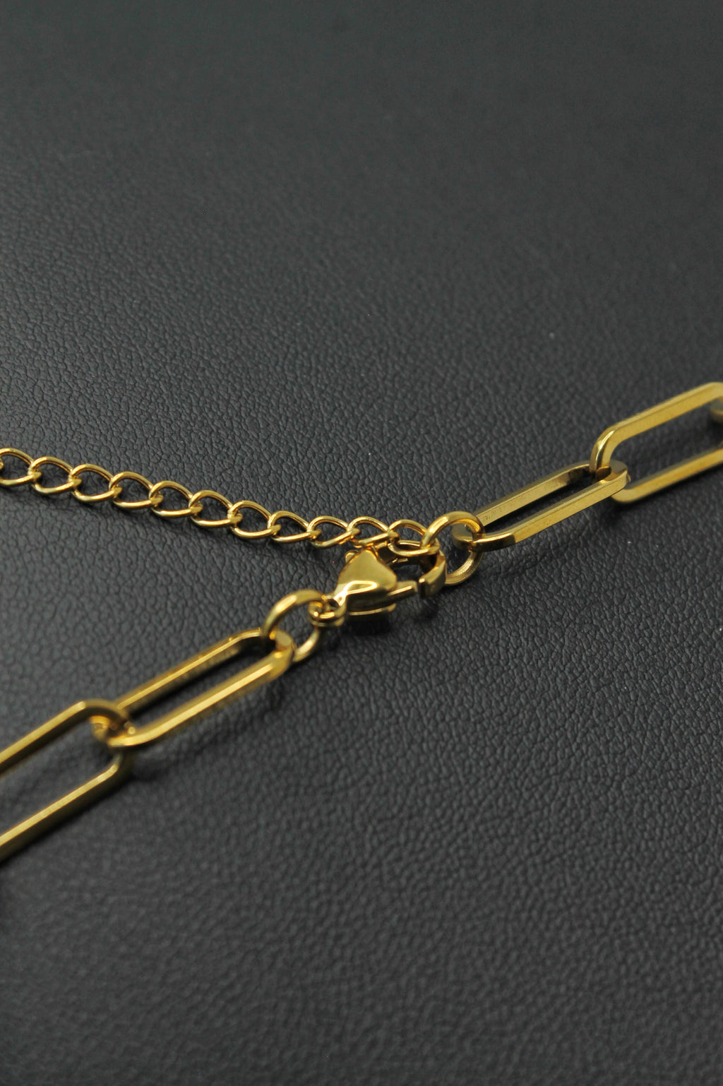 Close-up of a gold chain necklace with paperclip-style links and a lobster clasp, resting on a dark textured surface.