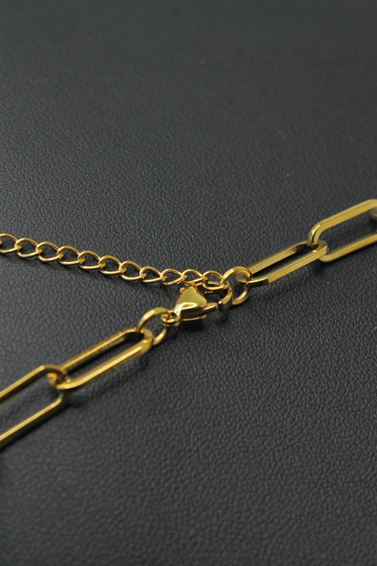 Close-up of a gold chain necklace with paperclip-style links and a lobster clasp, resting on a dark textured surface.