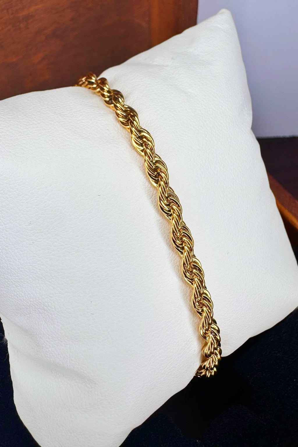 A gold-colored twisted rope chain bracelet displayed on a white fabric jewelry pillow.