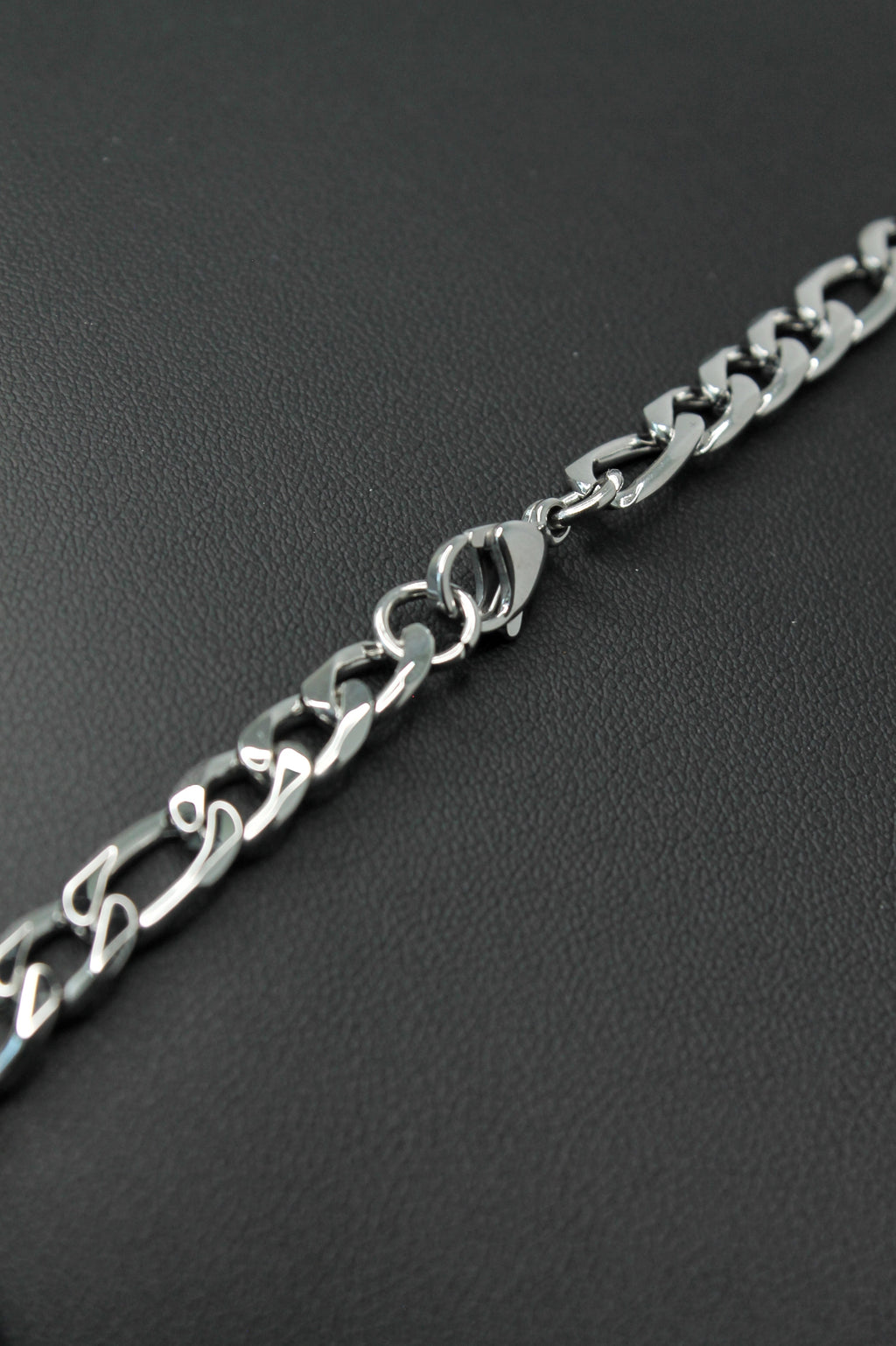 A close-up of a silver-toned metal Figaro chain bracelet with a lobster clasp, laid diagonally on a dark textured surface.