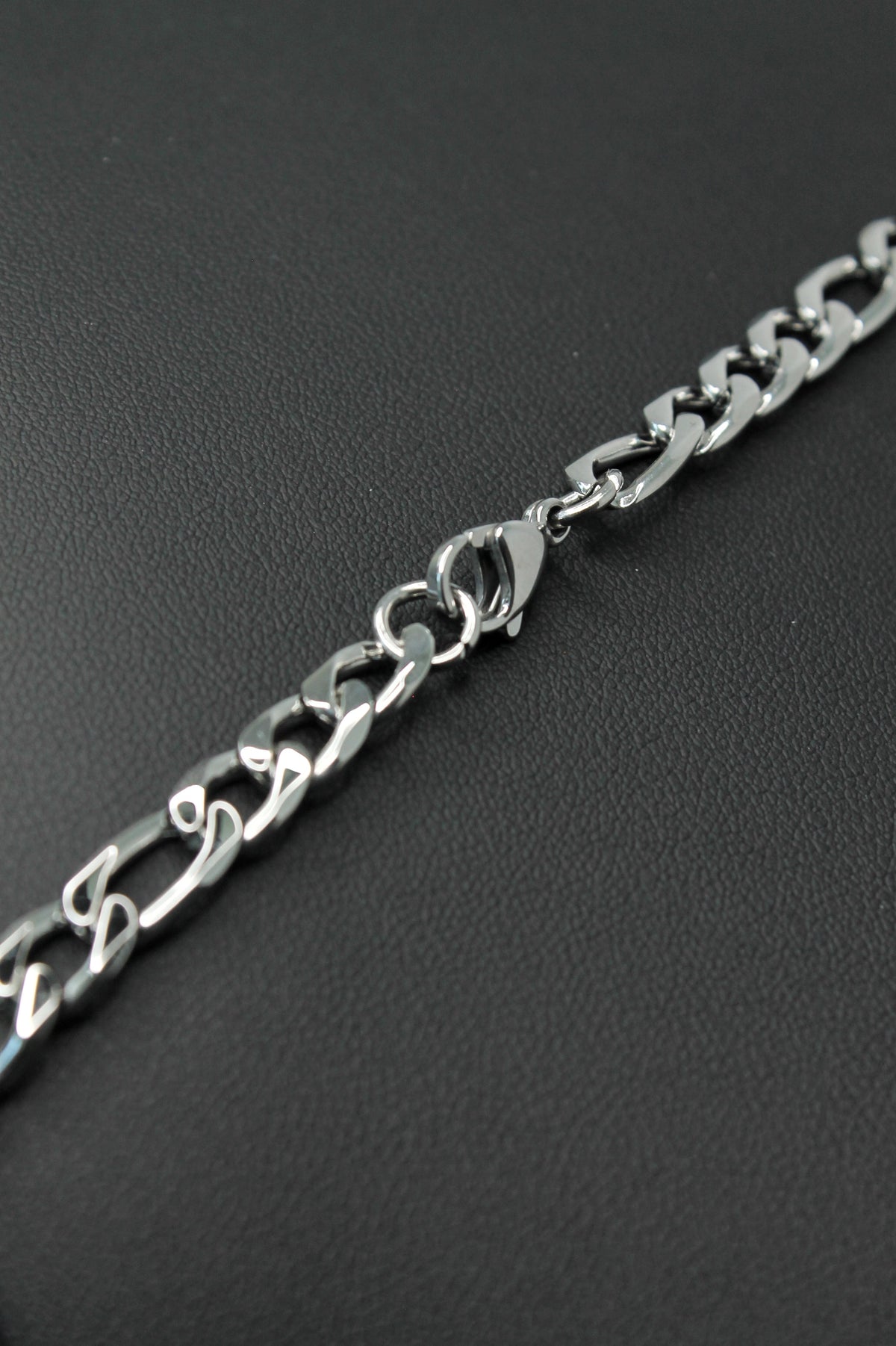 A close-up of a silver-toned metal Figaro chain bracelet with a lobster clasp, laid diagonally on a dark textured surface.