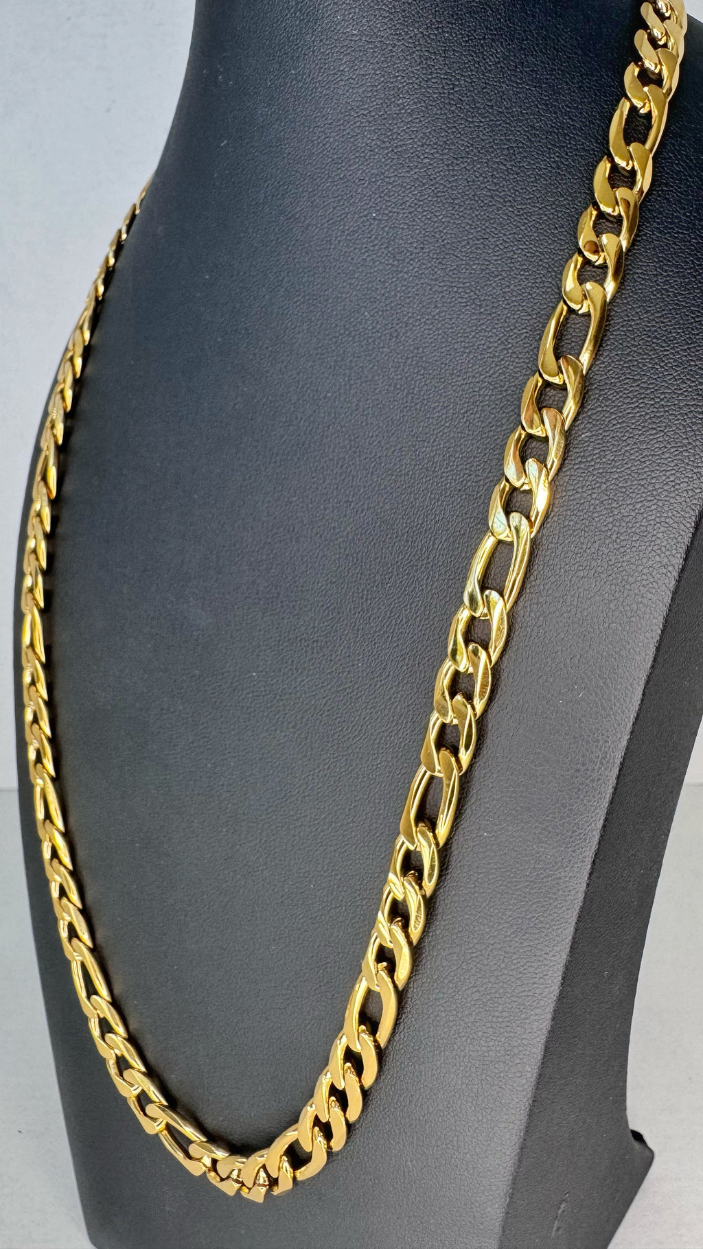 A chunky gold Figaro link chain necklace displayed on a black jewelry bust against a light gray background.