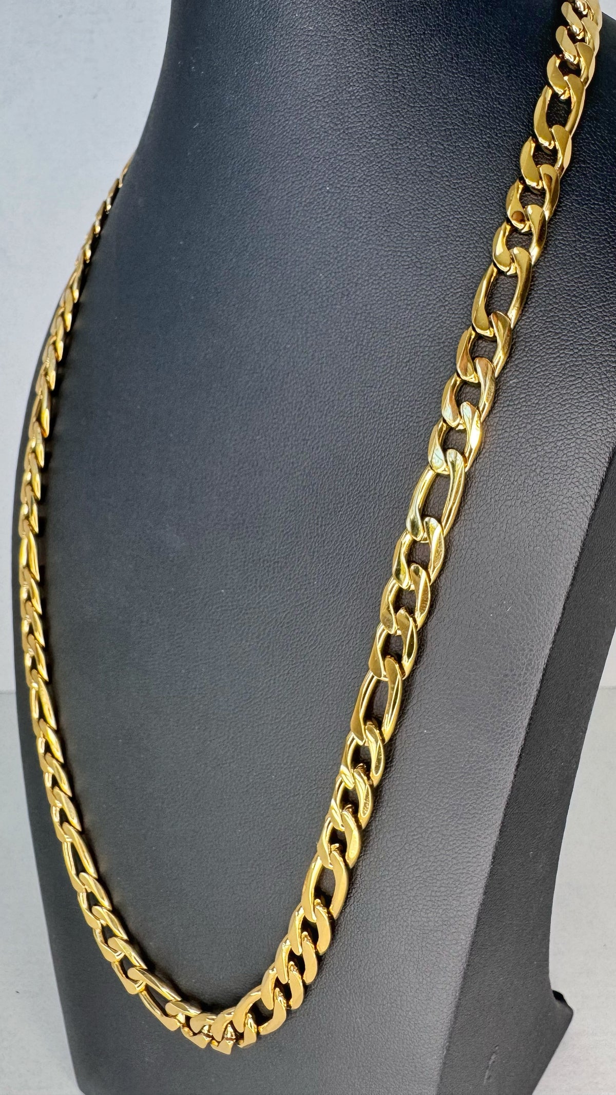 A chunky gold Figaro link chain necklace displayed on a black jewelry bust against a light gray background.