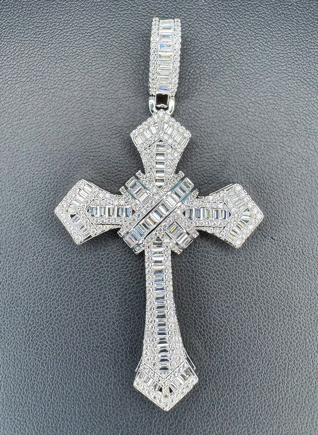 A silver-toned cross pendant encrusted with clear baguette and round-cut stones, displayed on a textured dark gray background.