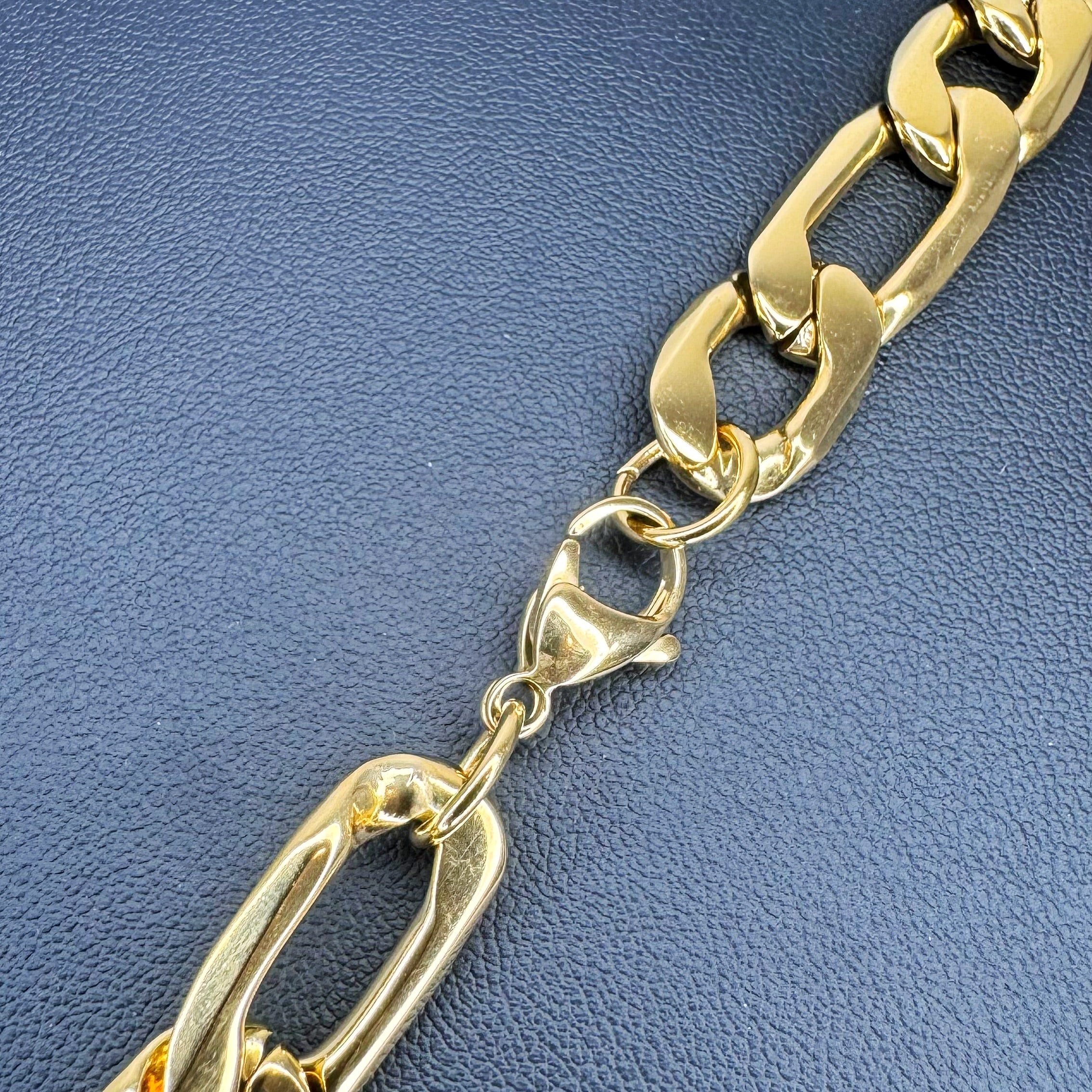 A close-up of a polished gold-tone chunky Figaro chain link bracelet on a textured blue background.