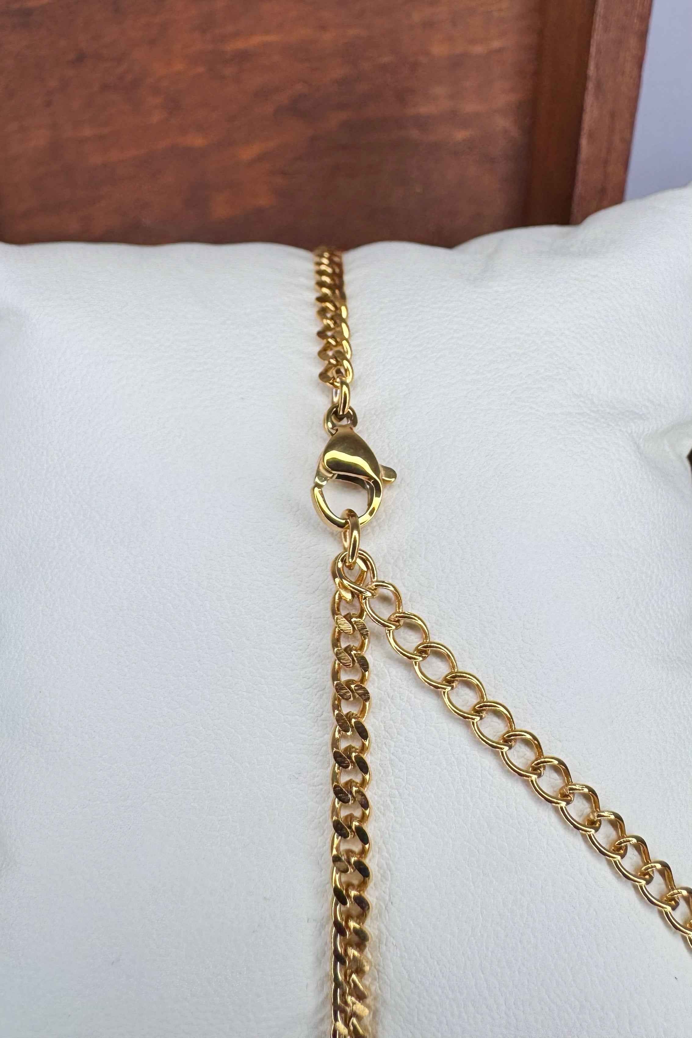 A close-up of a gold-tone Curb chain bracelet with a lobster clasp and chain extender, resting on a white display cushion.