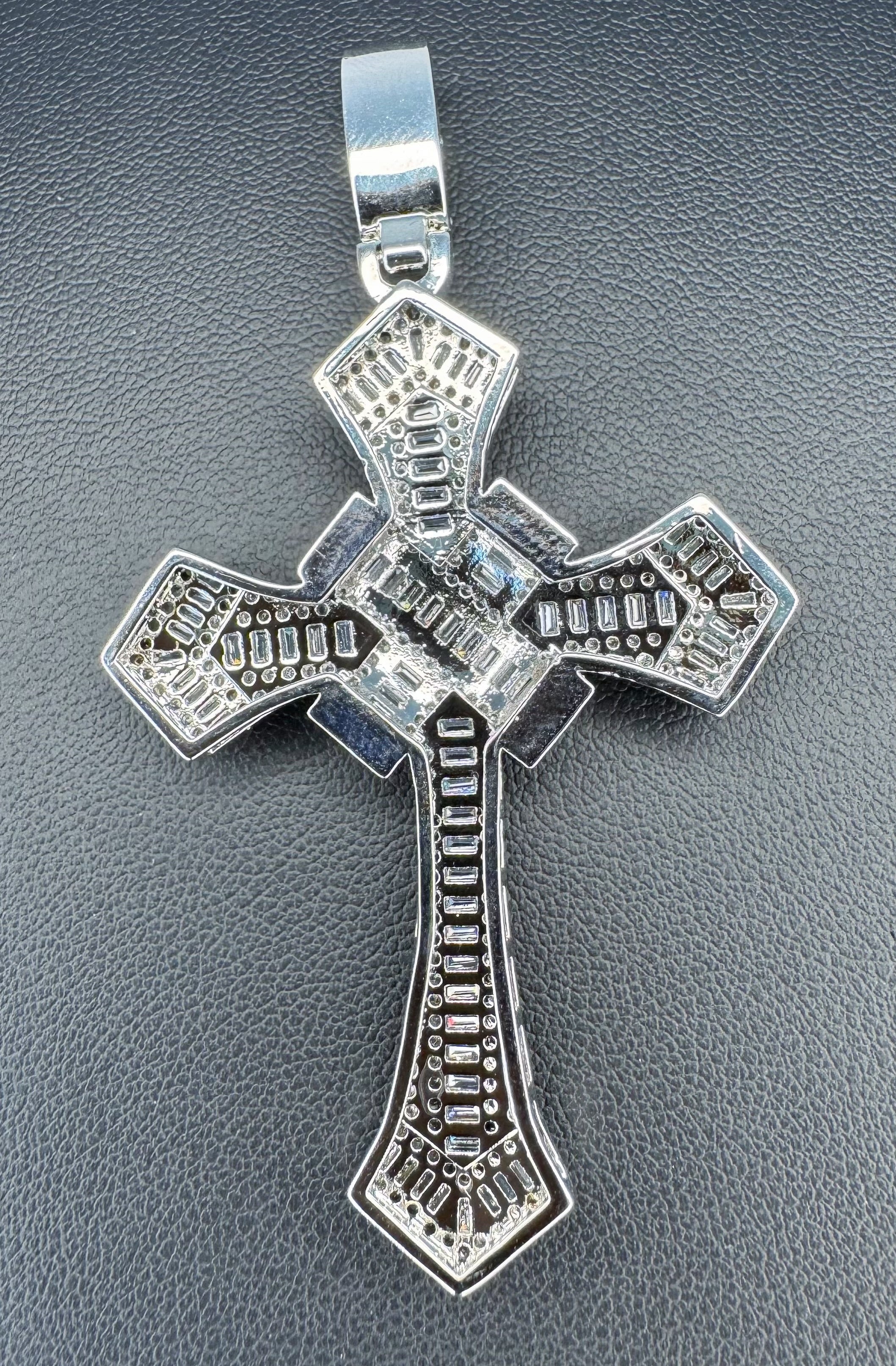 A silver cross pendant with intricate geometric filigree cutouts and a central clear gemstone, displayed on a textured dark gray background.
