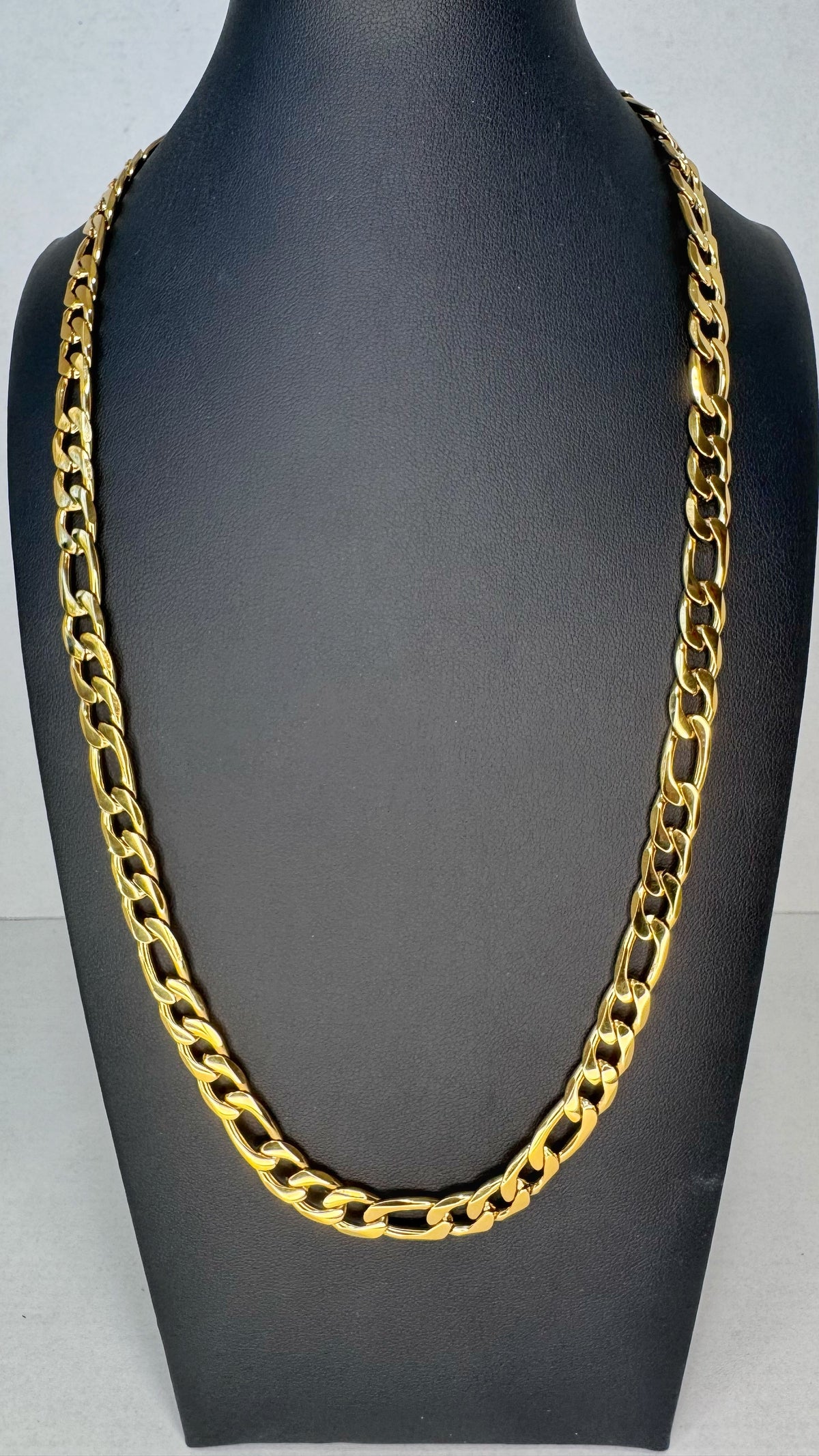 A chunky gold Figaro link chain necklace displayed on a black mannequin bust against a light gray background.