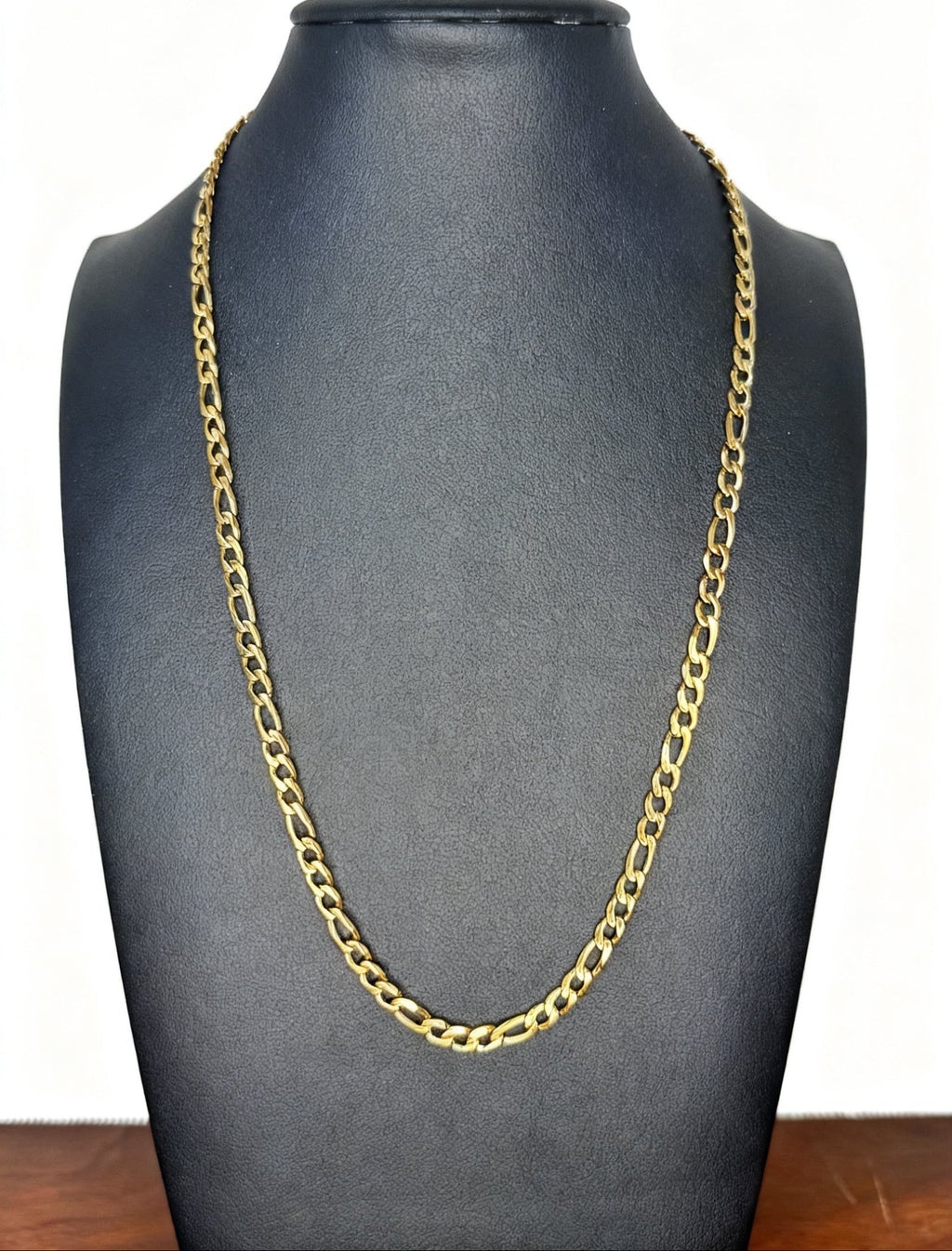 A gold-colored Figaro link chain necklace displayed on a matte black jewelry bust against a white background.