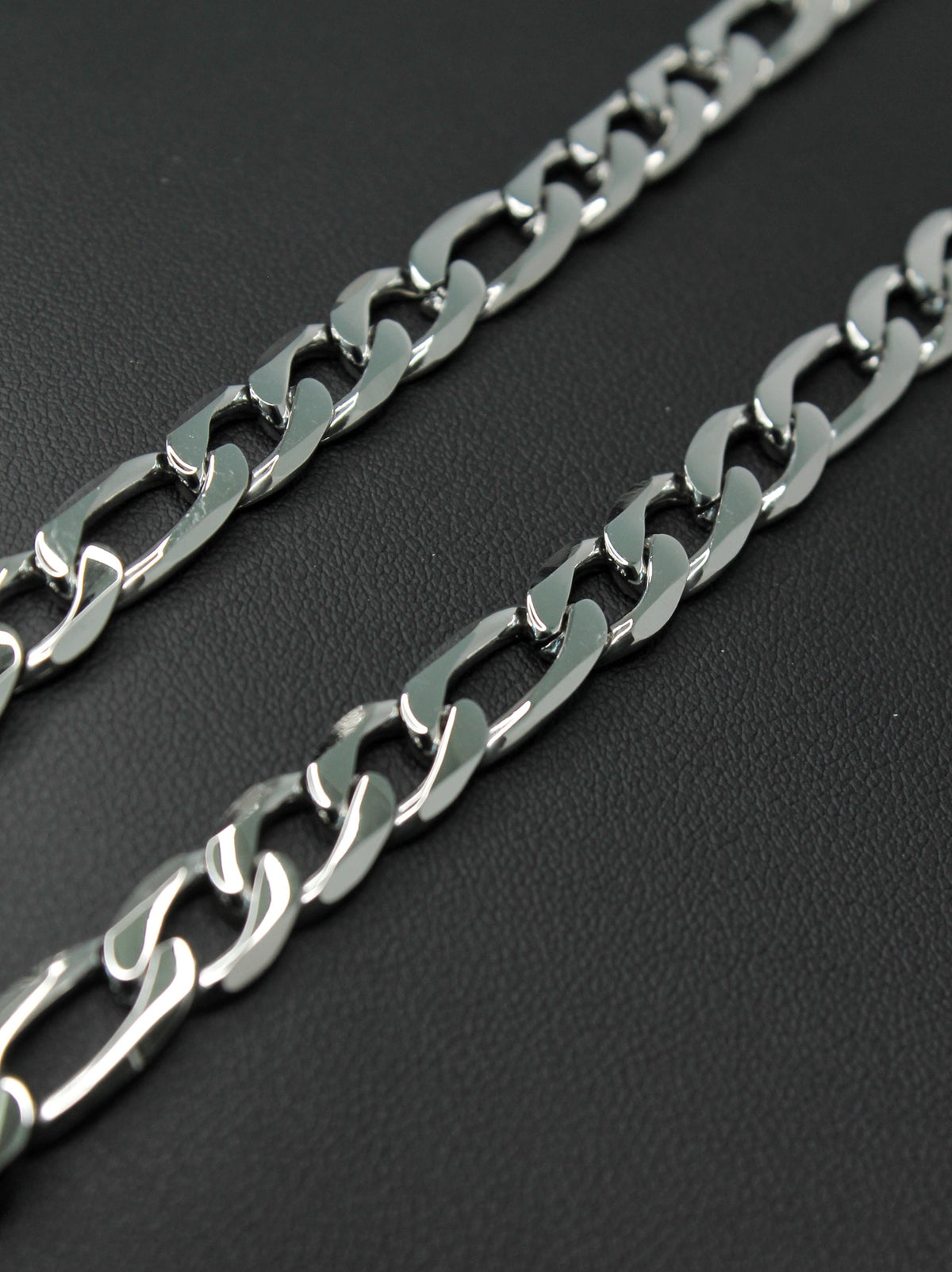 A close-up of a polished silver-tone metal Figaro chain with large interlocking links, laid diagonally on a dark textured background.