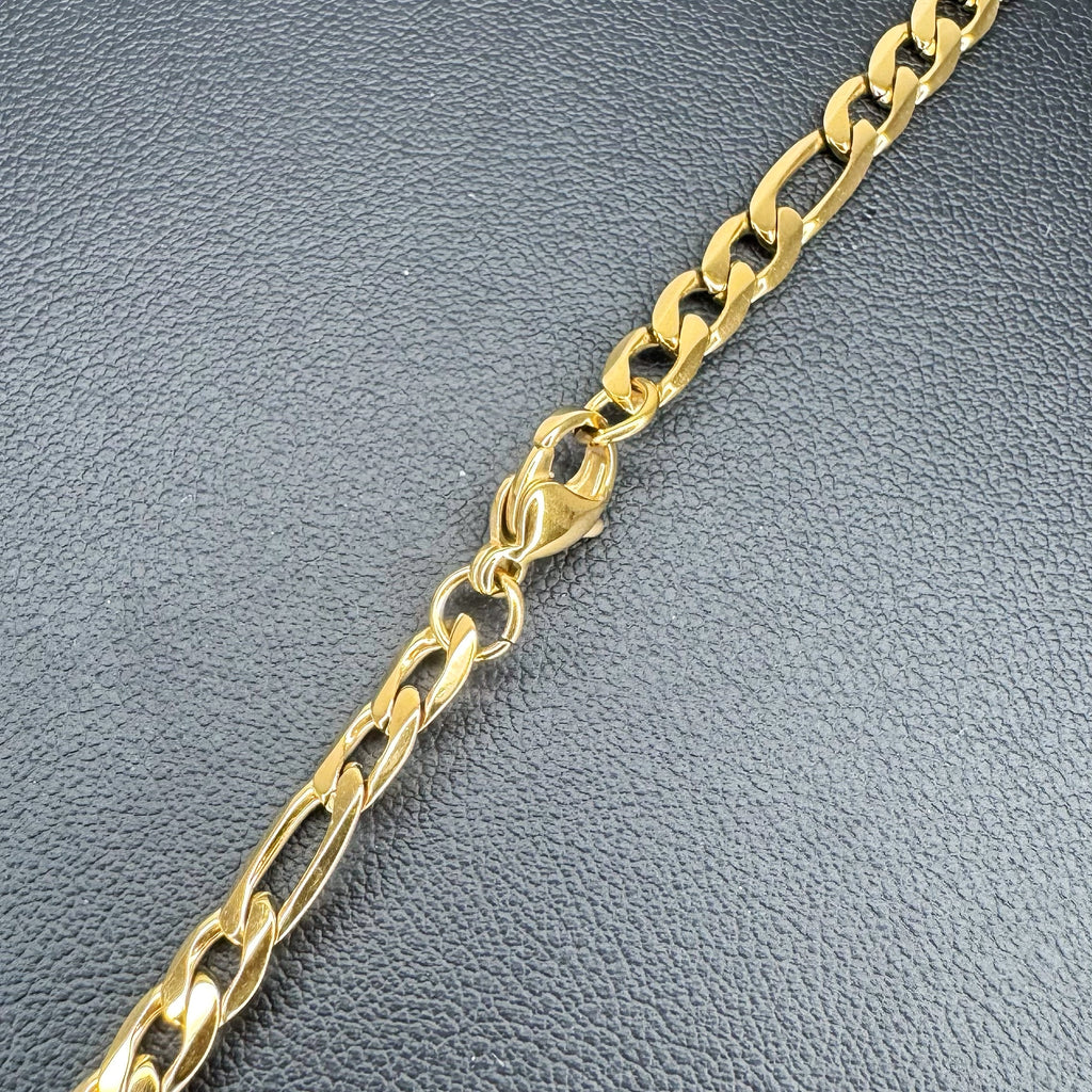 A close-up of a gold-toned Figaro link metal chain with a lobster clasp, laid diagonally on a textured dark gray surface.