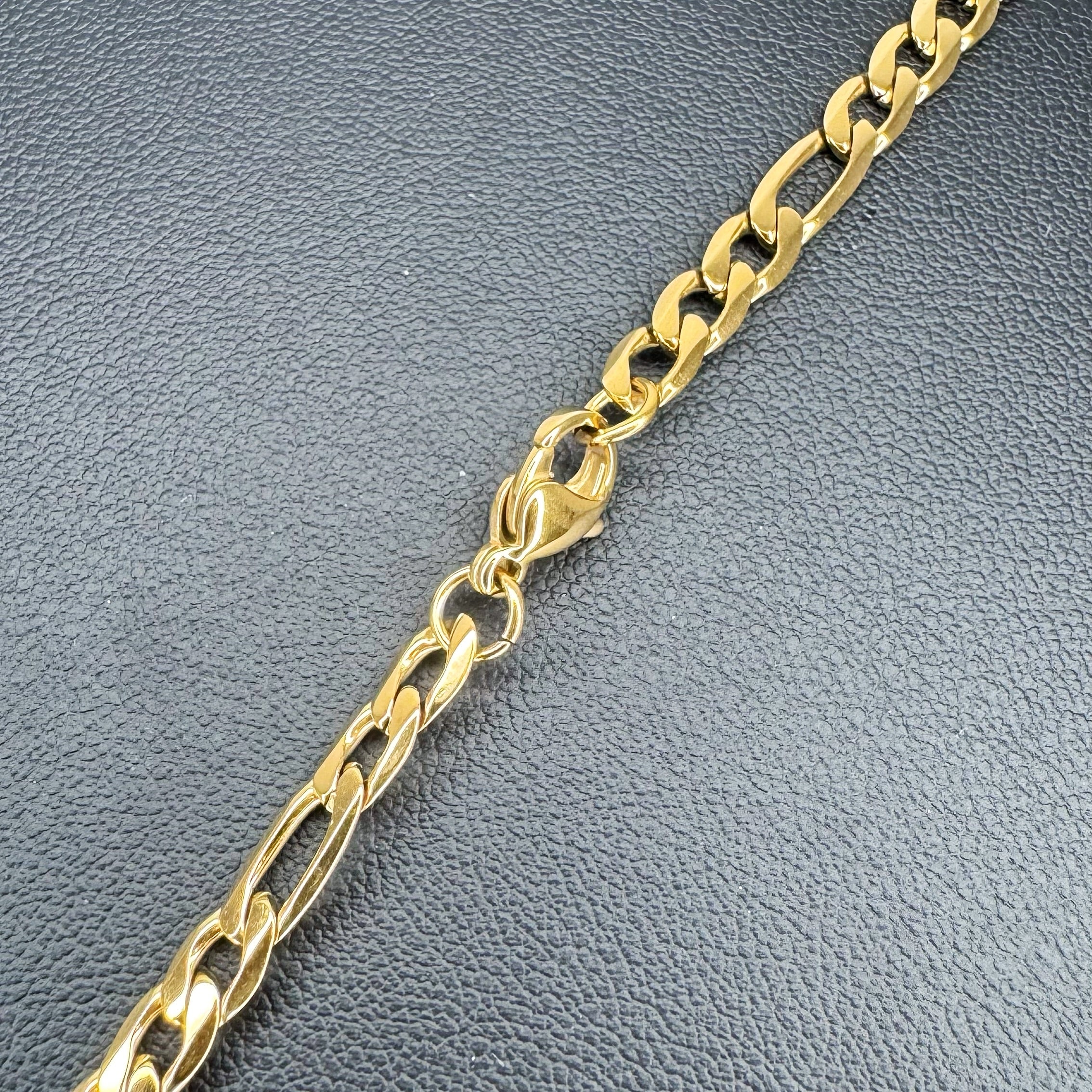 A close-up of a gold-toned Figaro link metal chain with a lobster clasp, laid diagonally on a textured dark gray surface.