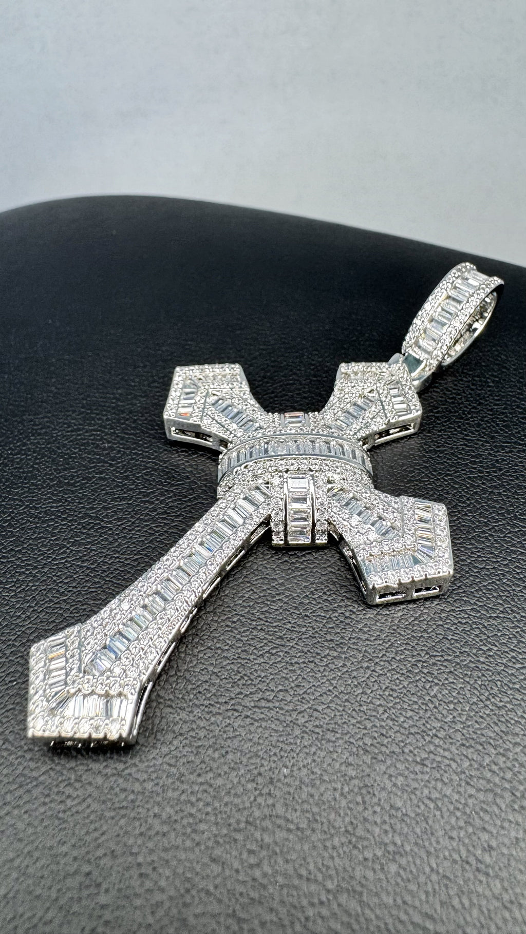 A large silver cross pendant encrusted with clear baguette and round-cut stones, displayed on a textured black surface.