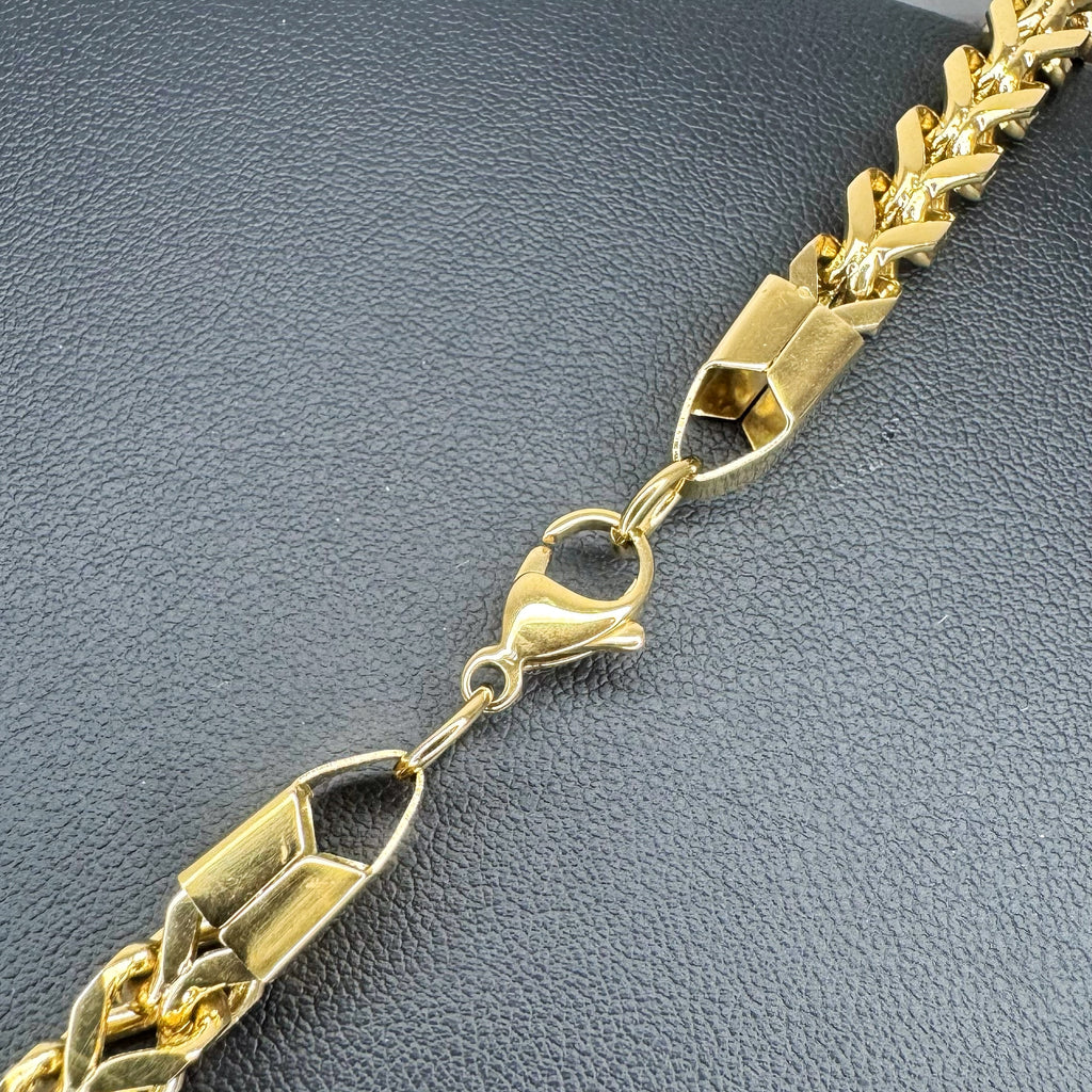 A close-up of a gold-toned Franco chain link bracelet with a lobster clasp, laid diagonally on a textured dark gray surface.