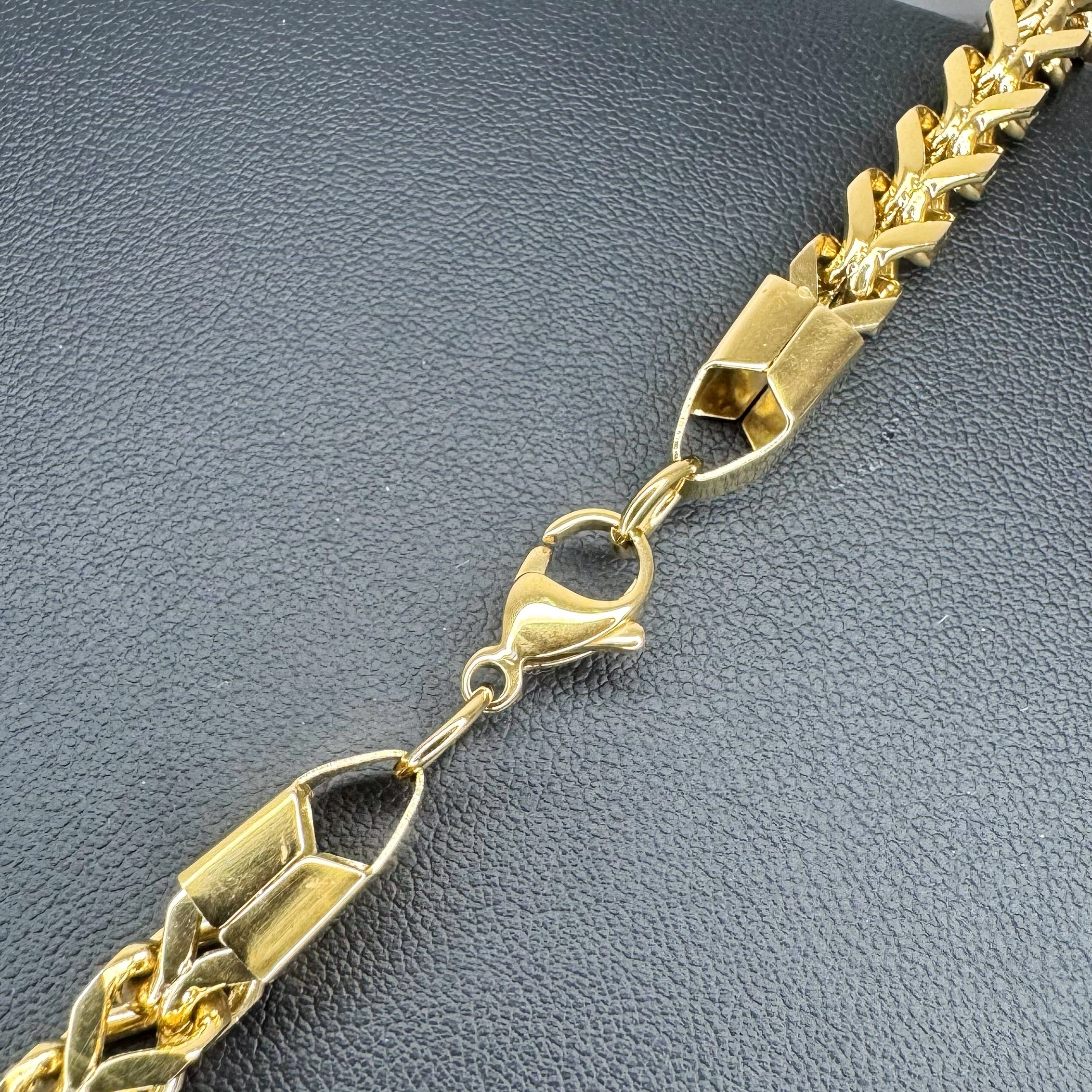 A close-up of a gold-toned Franco chain link bracelet with a lobster clasp, laid diagonally on a textured dark gray surface.
