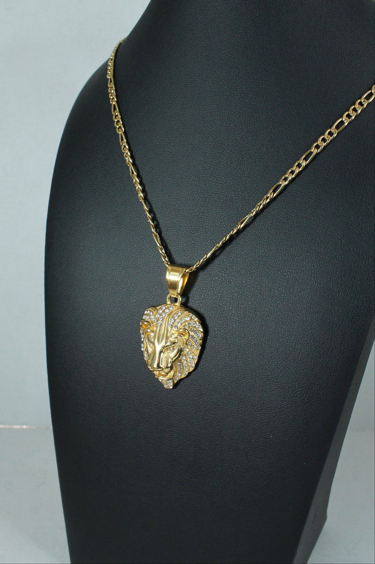 A gold chain necklace with a textured lion head pendant, displayed on a black mannequin bust against a light gray background.